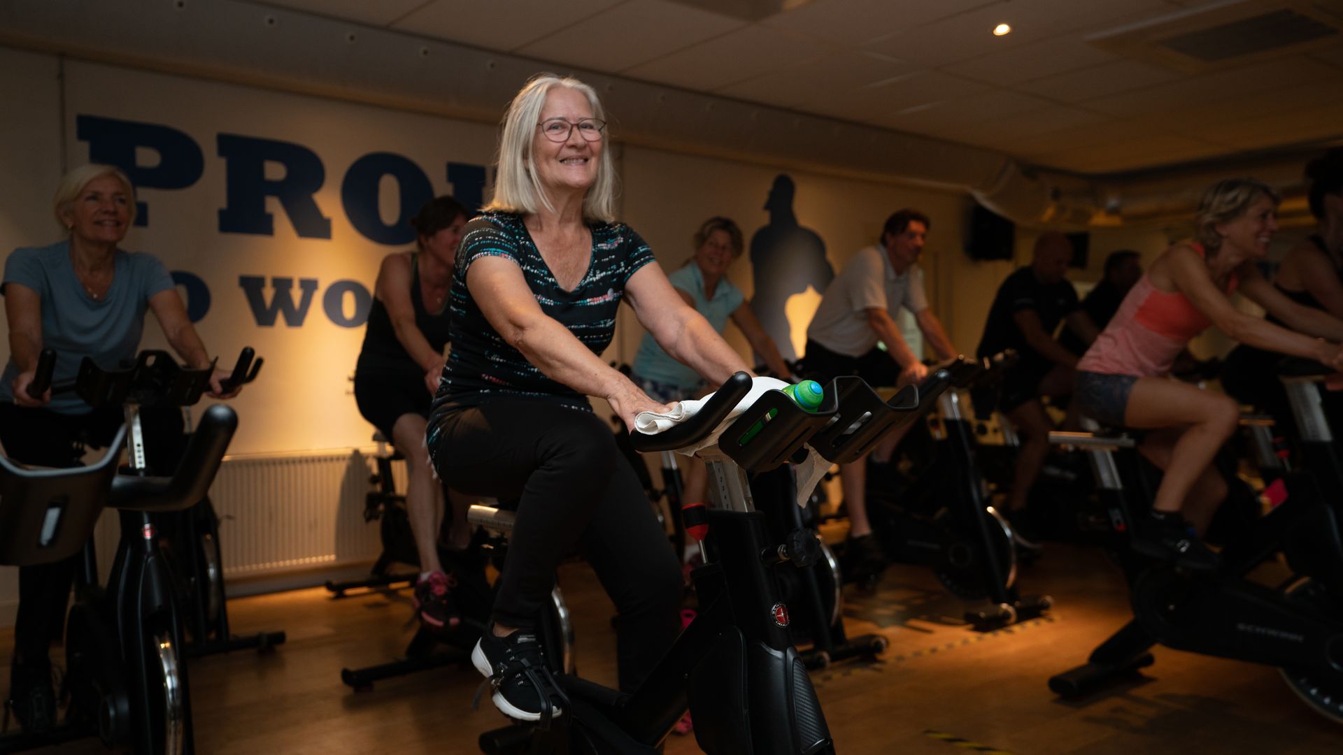 Een groep mensen fietst binnen. Een glimlachende vrouw in het midden leidt de les.