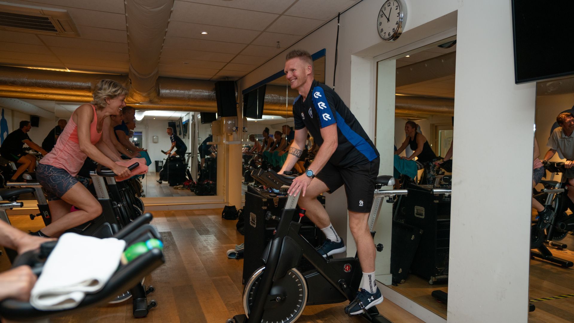 Een glimlachende spinning-instructeur begeleidt fietsers in een goed verlichte studio met spiegels.