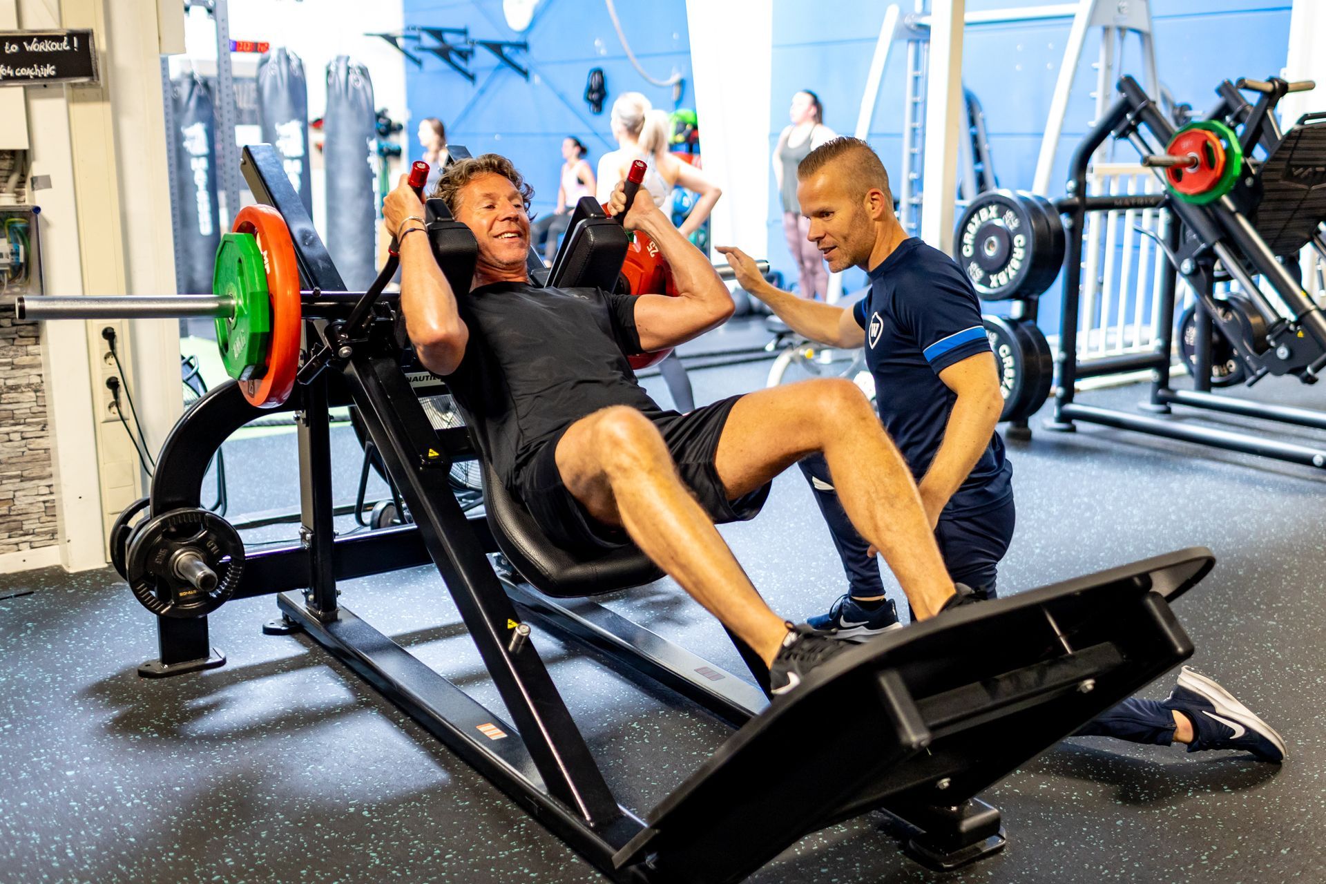Man gebruikt een leg press-apparaat in de sportschool, geholpen door een trainer. Ze lachen allebei.