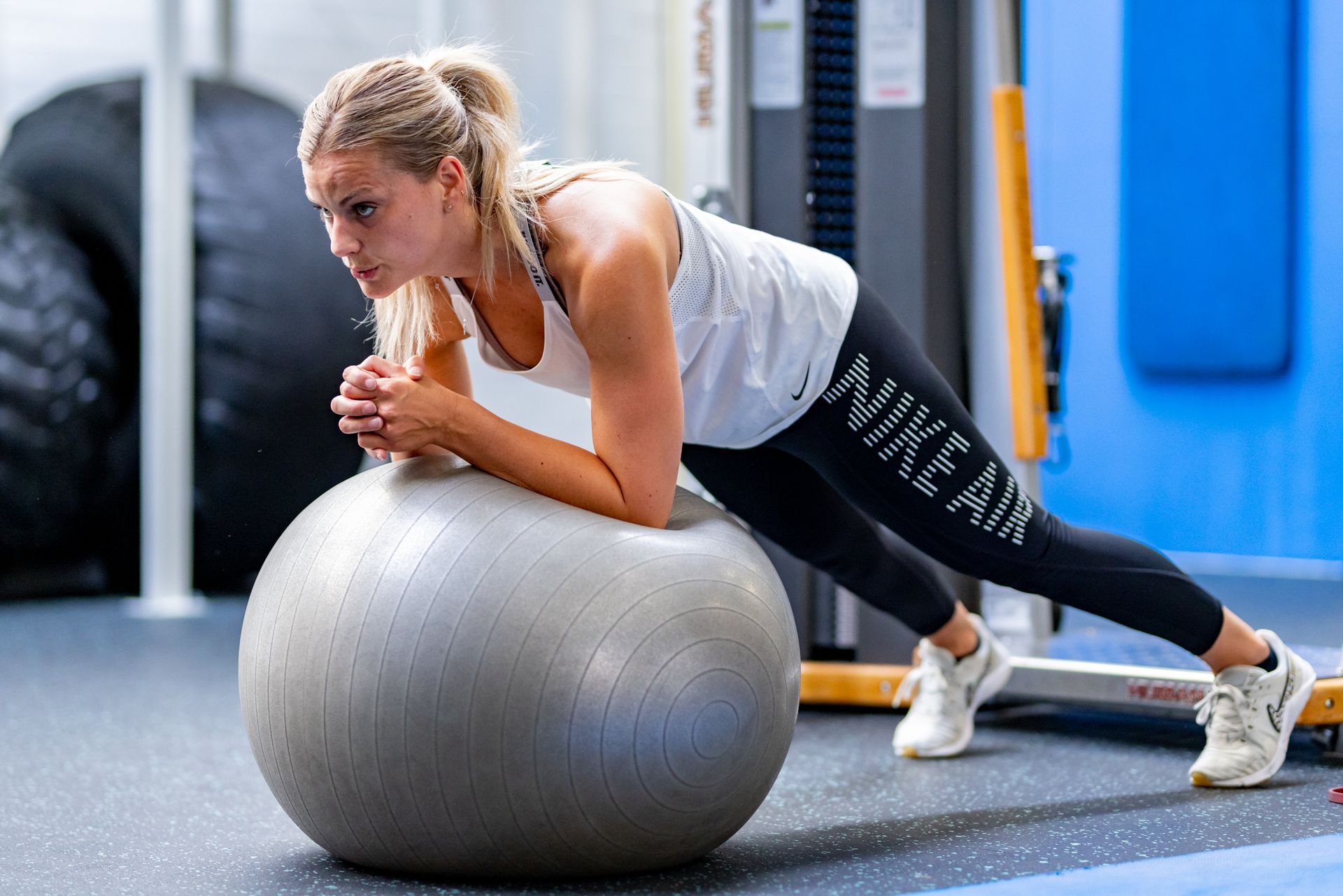 Een vrouw doet een plank op een fitnessbal in de sportschool. Ze draagt Nike-kleding en ziet er geconcentreerd uit.