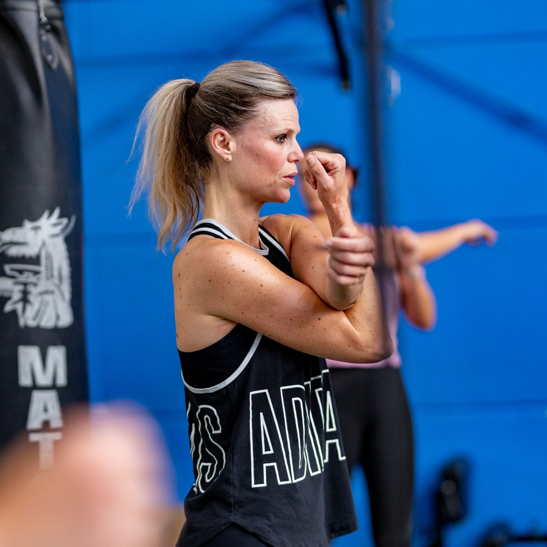Een vrouw in de sportschool strekt haar arm uit; ze draagt een zwart tanktopje met witte letters.