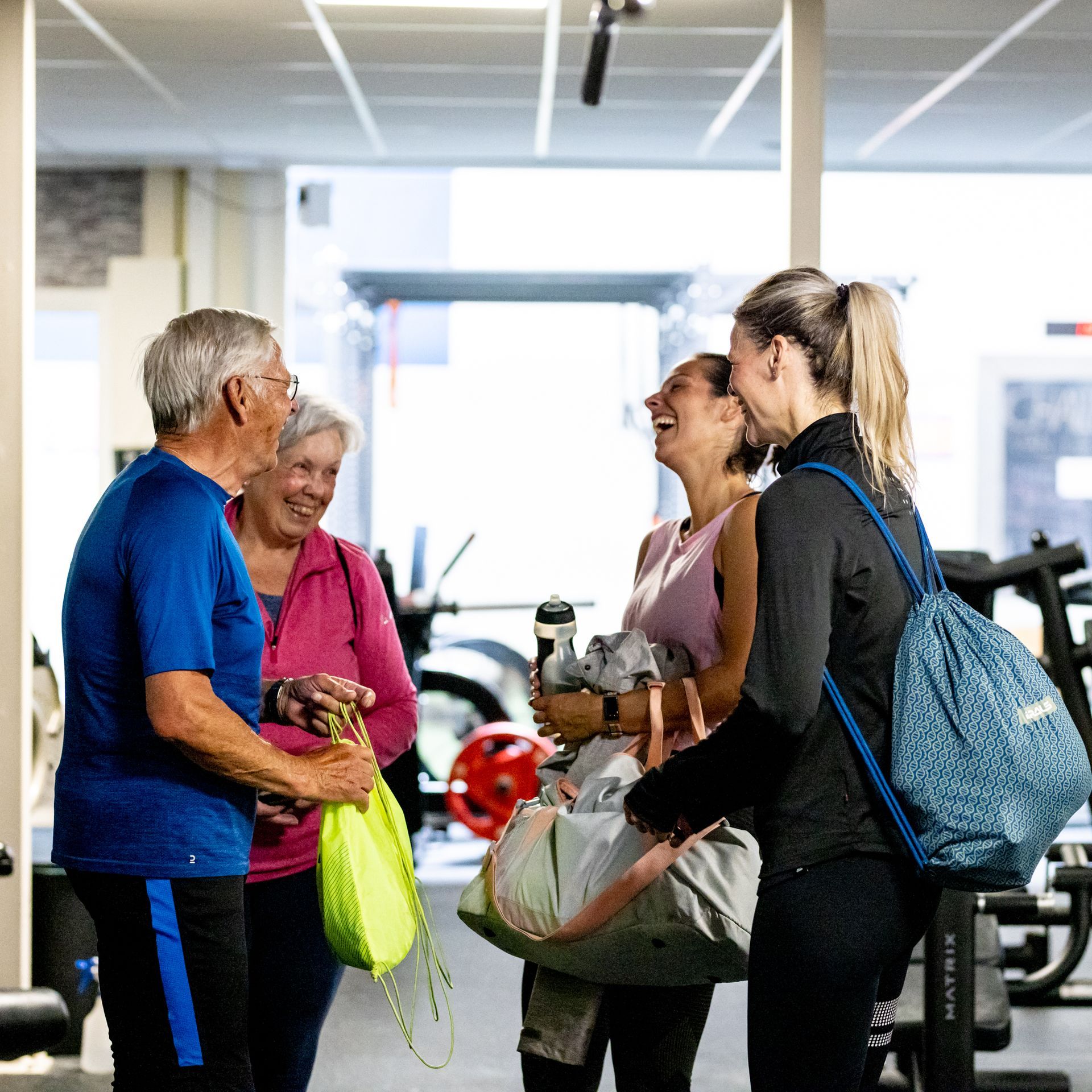 Vier mensen lachen samen in een sportschool. Twee oudere volwassenen, twee jongere vrouwen, lachend, met tassen in hun handen.