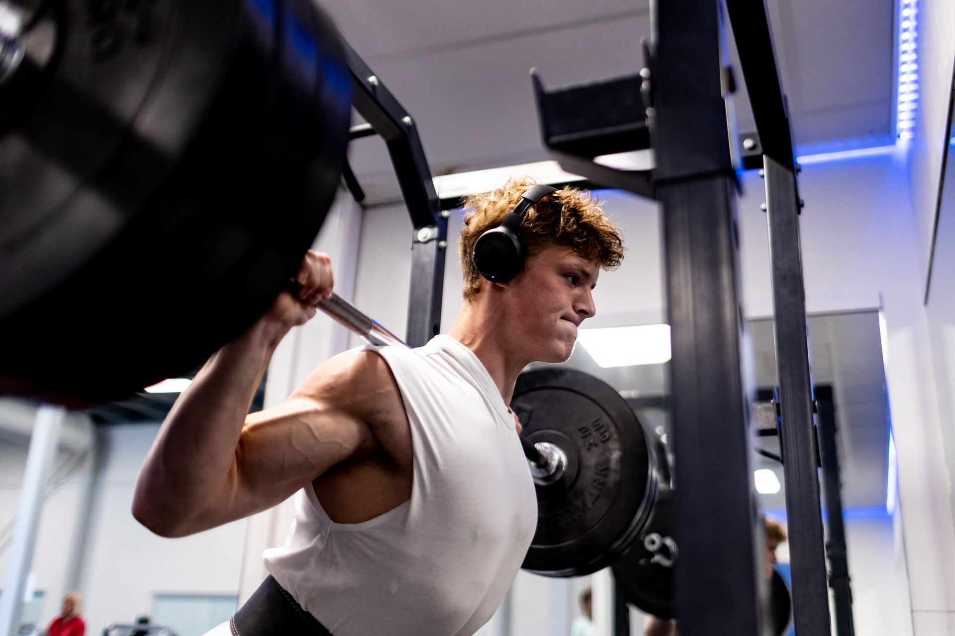 Man hurkt met halter in sportschool, draagt koptelefoon, geconcentreerde uitdrukking.