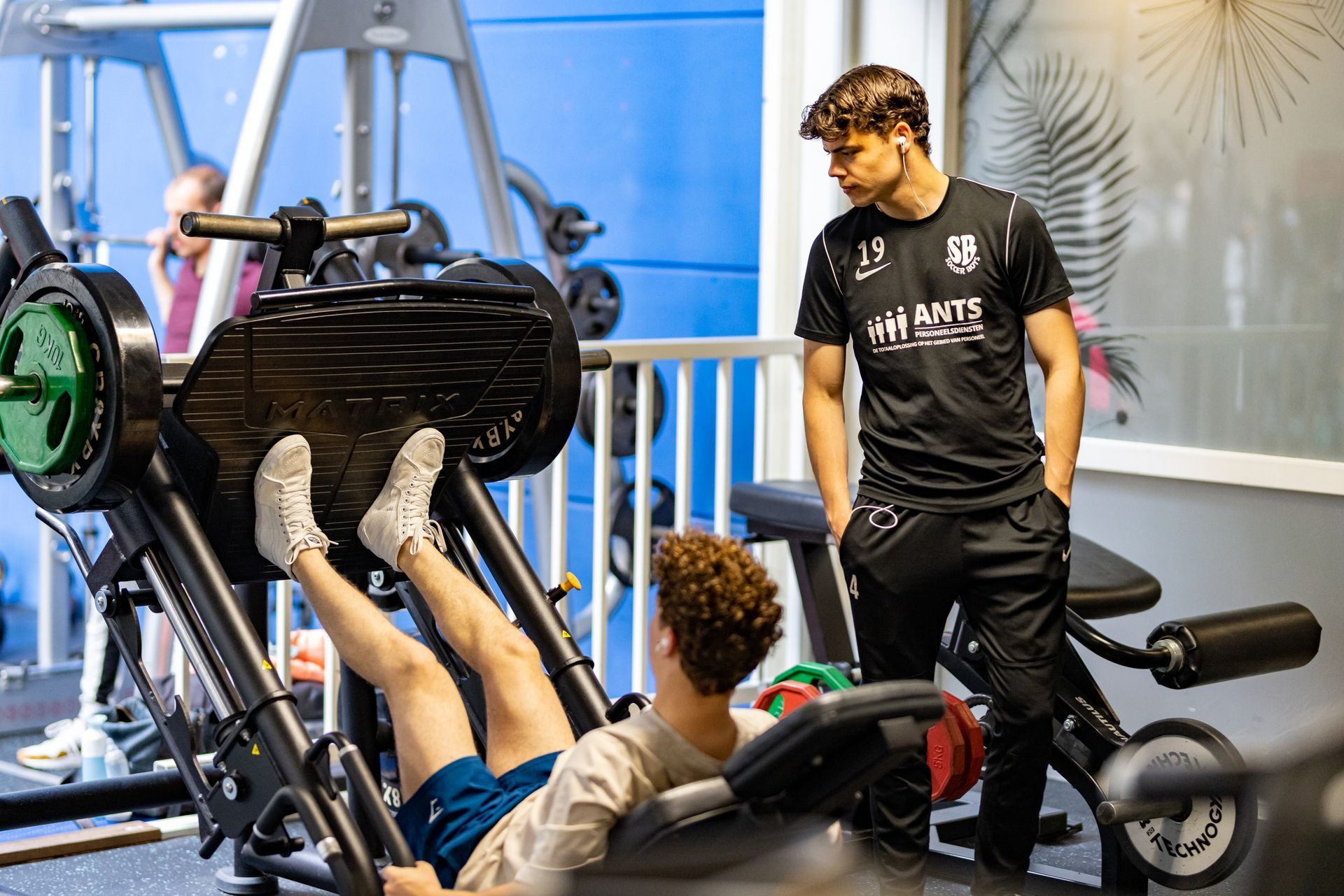 Twee mannen in een sportschool: de een doet een leg press, de ander kijkt toe en coacht.