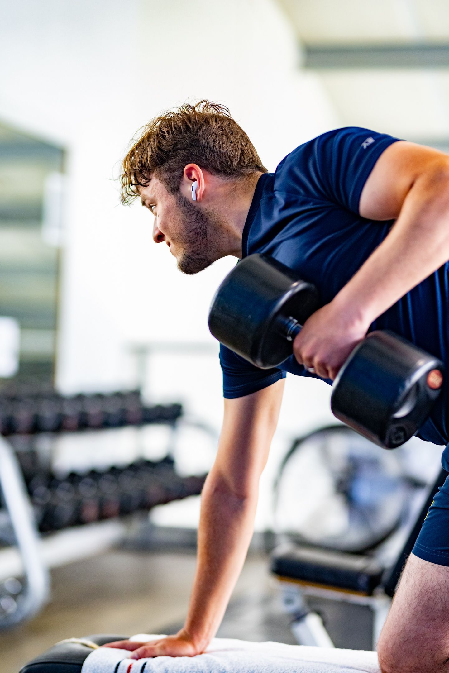 Man in de sportschool tilt een halter op. Hij draagt een blauw shirt en een korte broek, met gewichten op de achtergrond.