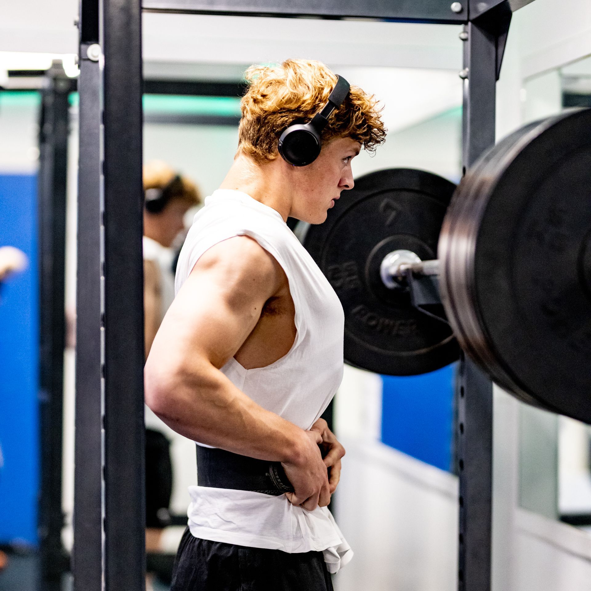 Jongeman met koptelefoon maakt zich klaar om gewichten te heffen in de sportschool. Zwarte halterstang en gewichtenrek zijn zichtbaar.