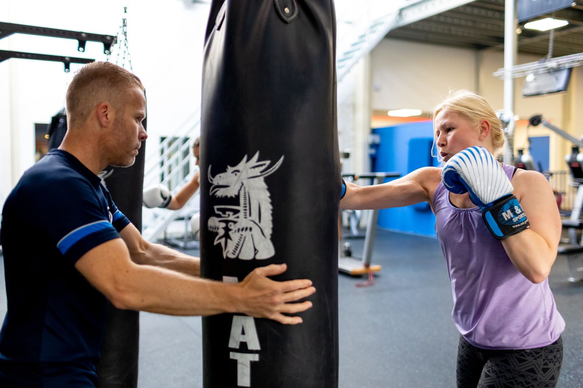 Een vrouw met bokshandschoenen slaat met een bokszak, terwijl een man haar traint in de sportschool.