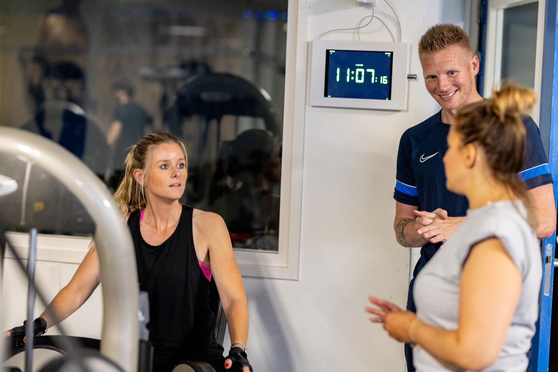 Drie mensen in een sportschool: een vrouw op een apparaat, twee mensen tegenover haar, allemaal lachend en pratend.
