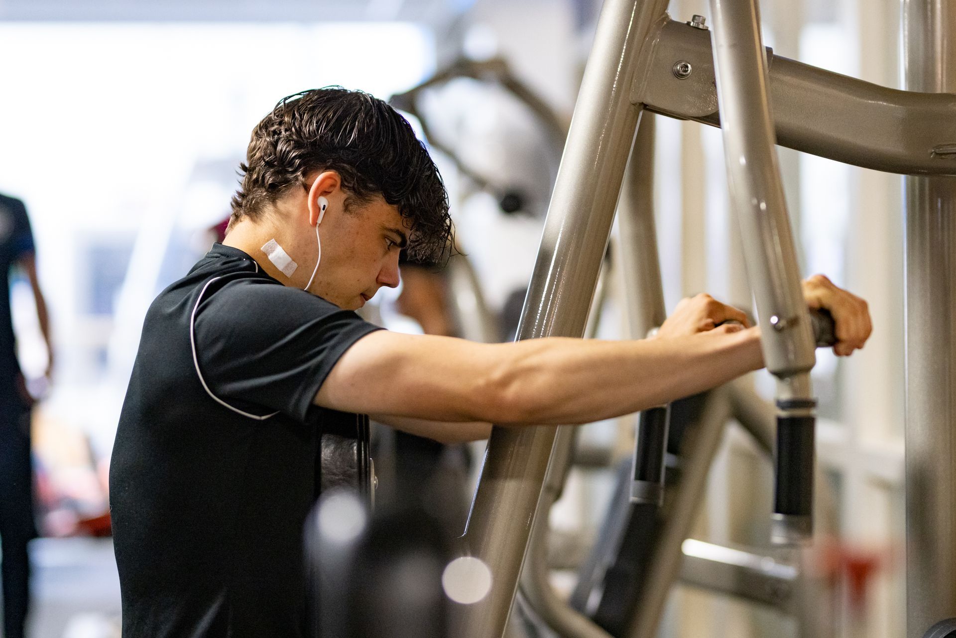 Persoon die in de sportschool een borstpersapparaat gebruikt, draagt een zwart shirt, oordopjes en een geconcentreerde gezichtsuitdrukking.