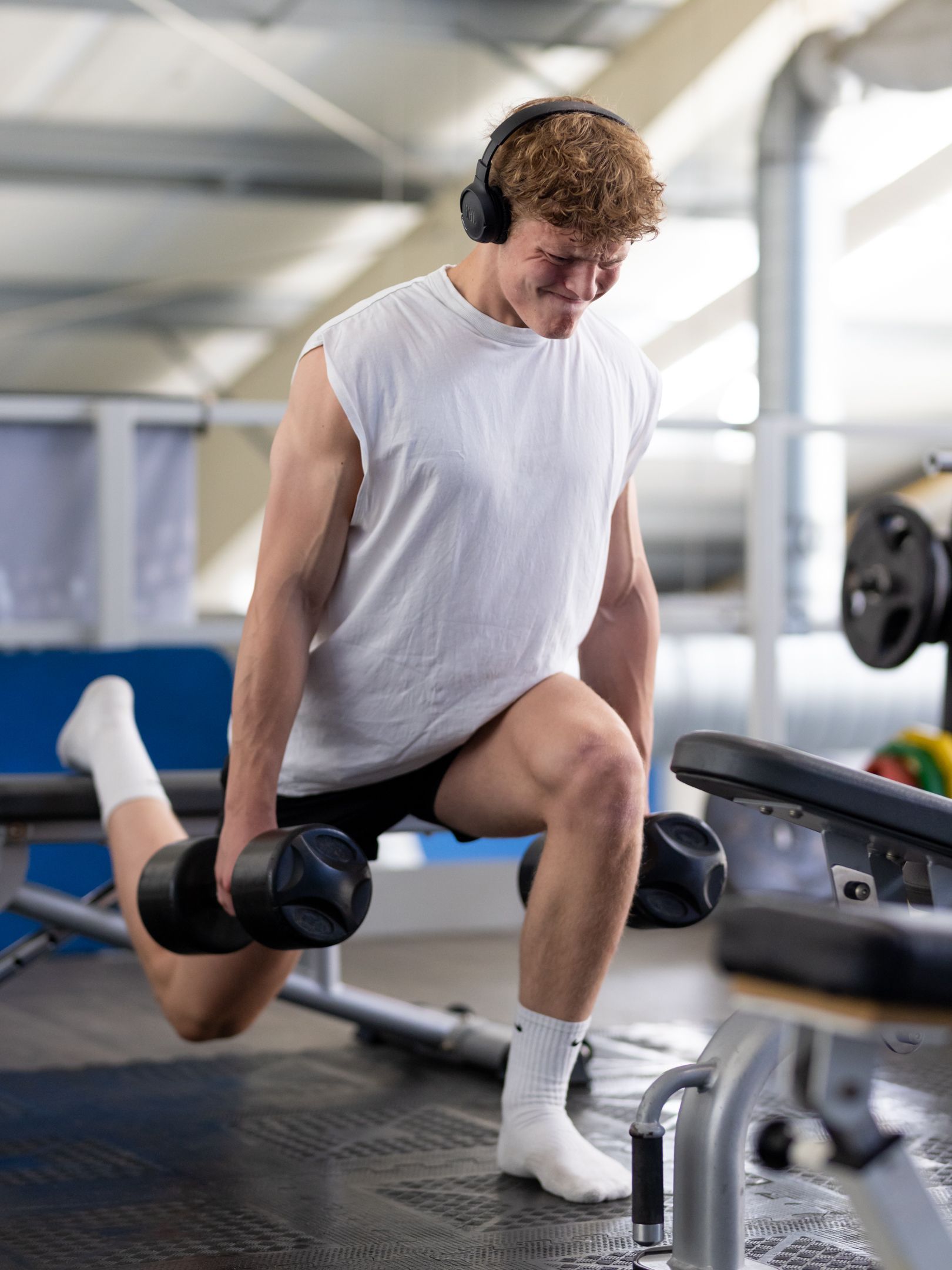 Man doet dumbbell lunges in de sportschool, draagt een koptelefoon, een wit shirt, sokken en een zwarte korte broek.