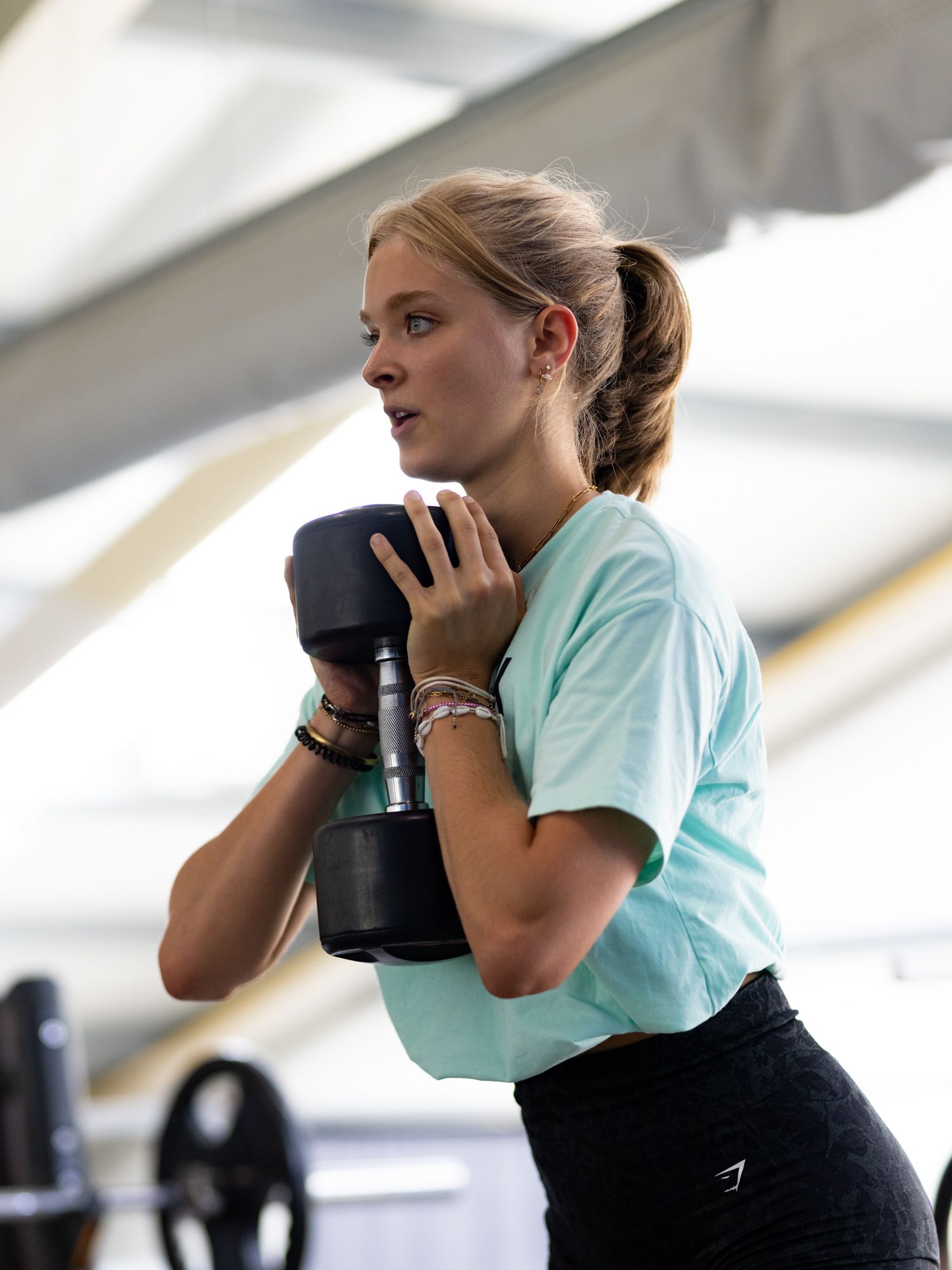 Een vrouw in een sportschool met een halter in haar handen, klaar om te hurken. Ze draagt een blauw shirt en een zwarte legging.