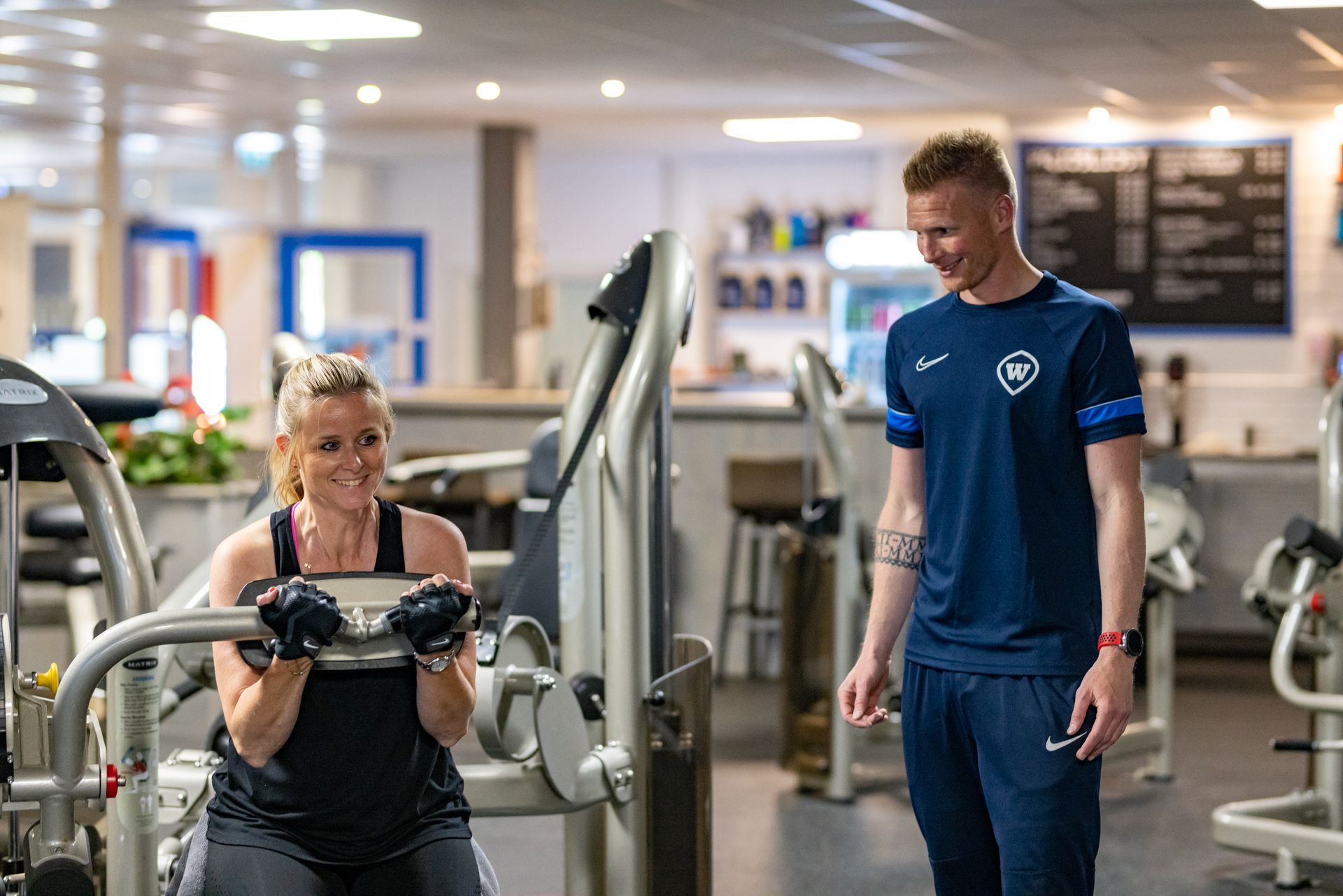Vrouw gebruikt een gewichtsapparaat, bijgestaan ​​door een personal trainer in een sportschool.