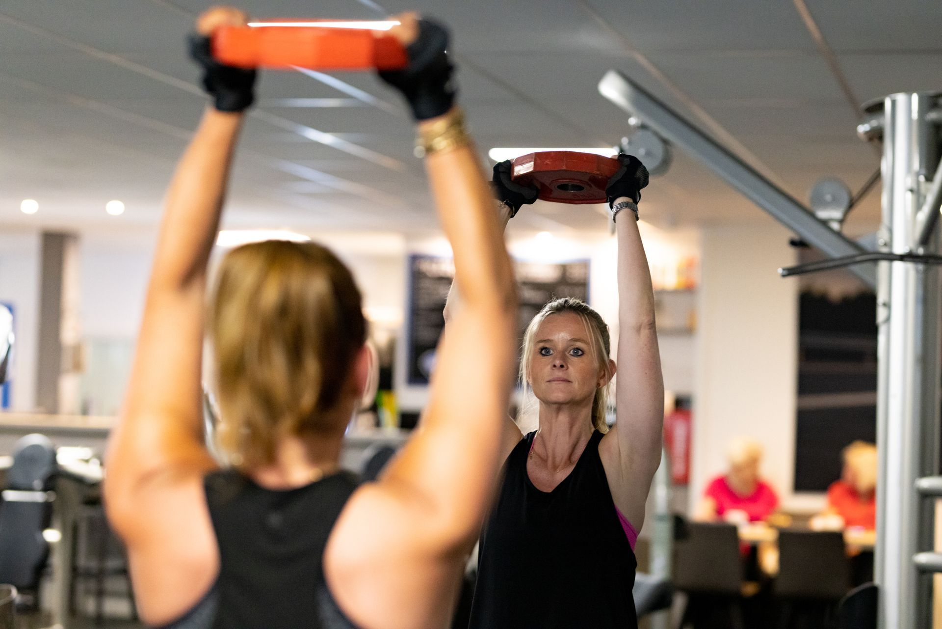Twee vrouwen tillen gewichten boven hun hoofd in een sportschool; één op de voorgrond, de ander op de achtergrond.