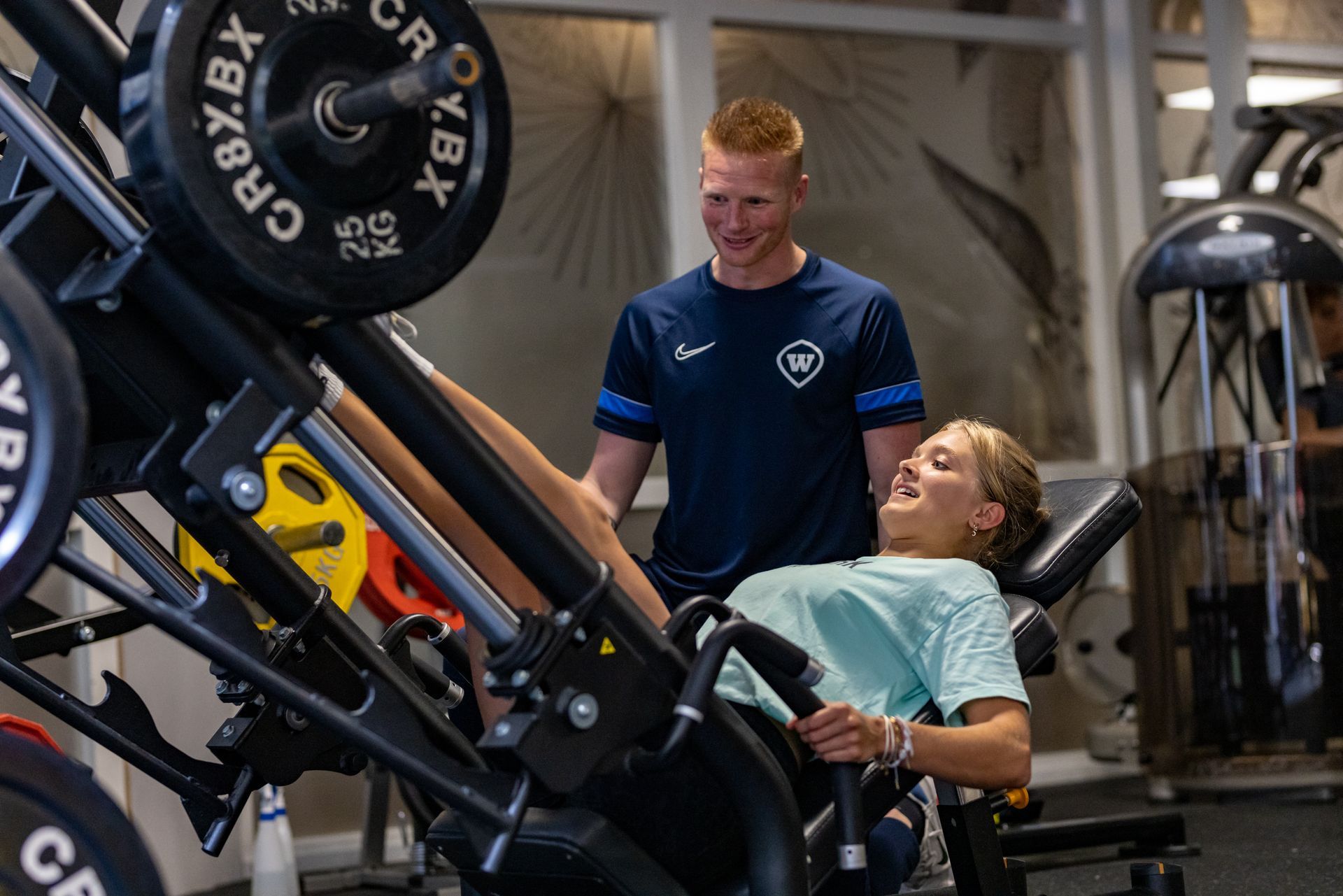 Een vrouw gebruikt een leg press-apparaat met een trainer in de sportschool. Ze lachen allebei.