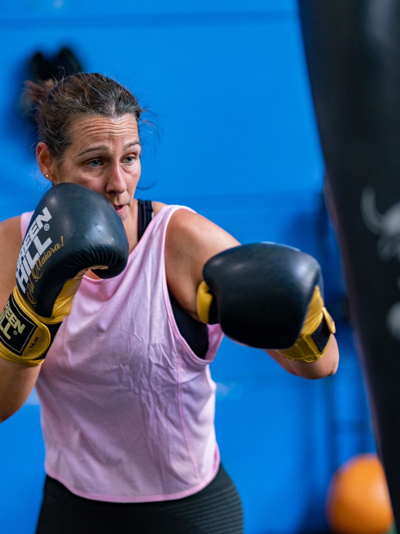 Vrouw in roze tanktop doet boksen met een boksbal en zwarte en gouden handschoenen in een blauwe sportschool.