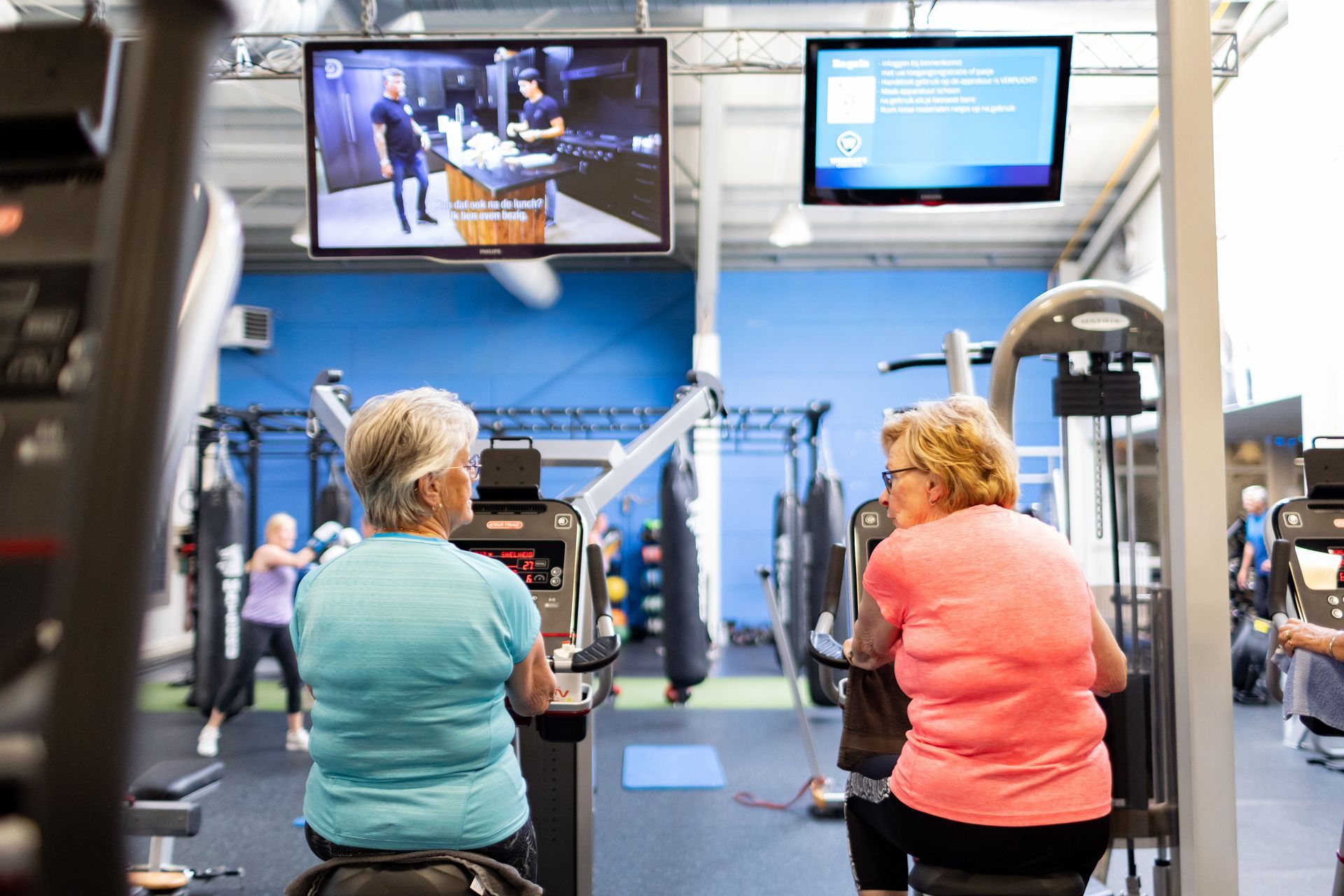 Twee oudere vrouwen trainen op apparaten in een sportschool en kijken tv. Anderen trainen op de achtergrond.
