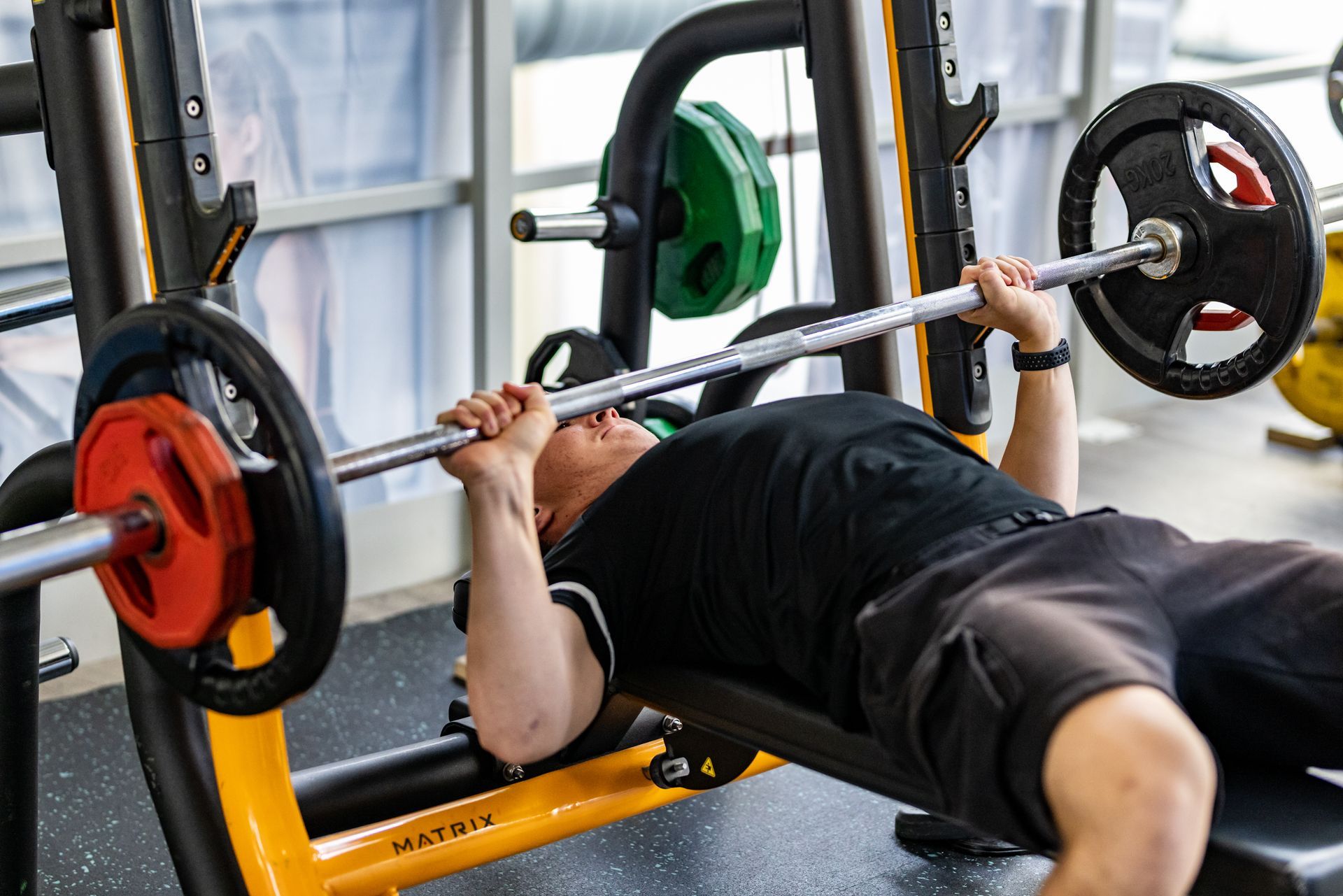 Man bankdrukt met halterstang in de sportschool. Oranje en groene gewichten zichtbaar.