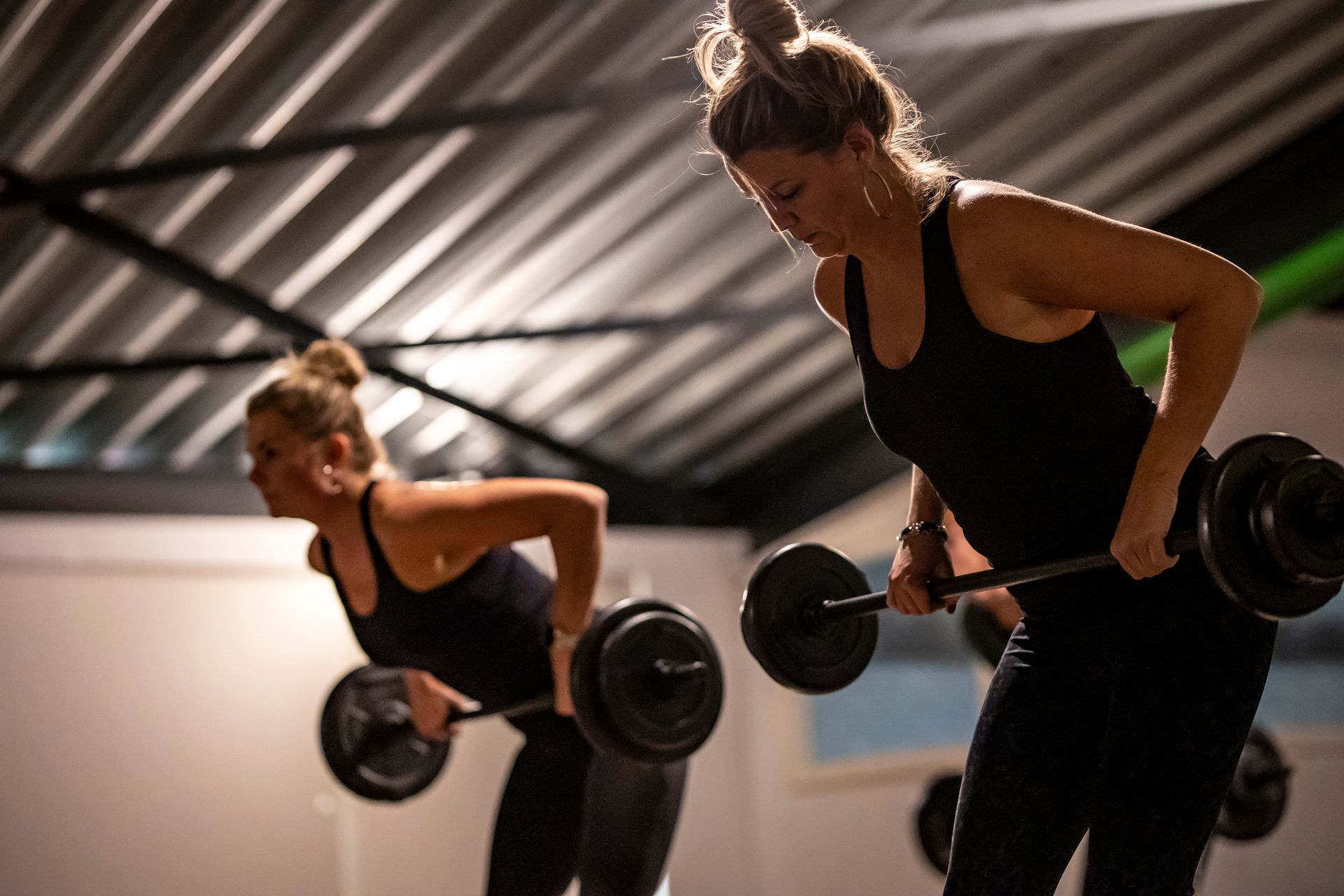 Twee vrouwen in een sportschool, voorovergebogen, tillen halters. Ze dragen zwarte tanktops en leggings.