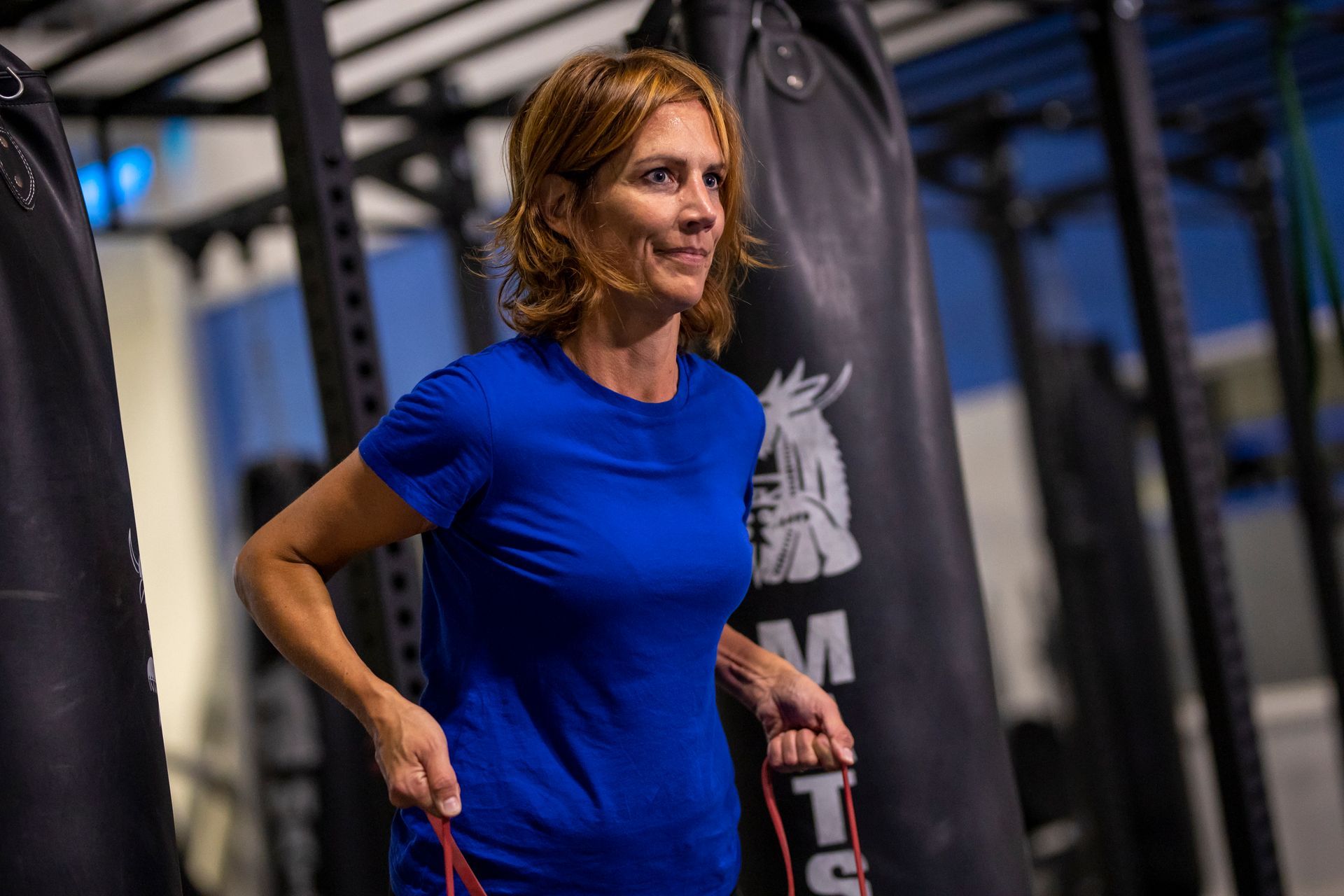 Vrouw in een blauw shirt traint met weerstandsbanden in een sportschool, vlakbij bokszakken.