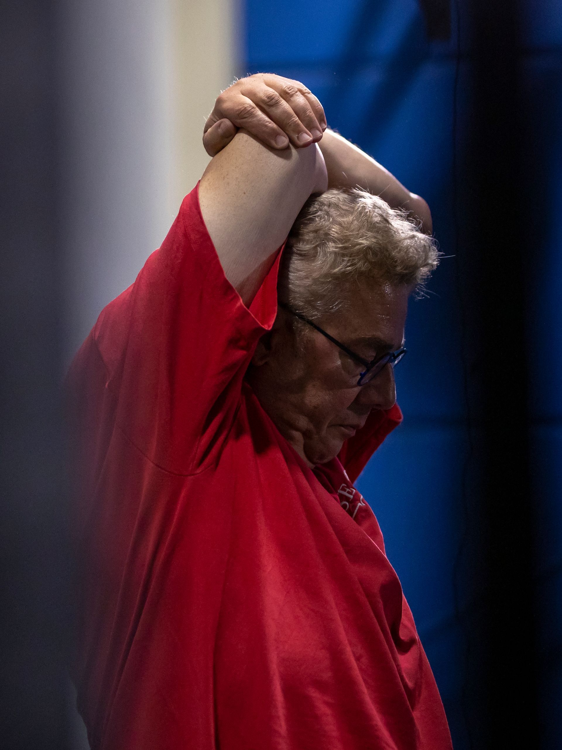 Persoon in rood shirt, armen achter het hoofd, naar beneden kijkend. Wit haar, bril, blauwe achtergrond.