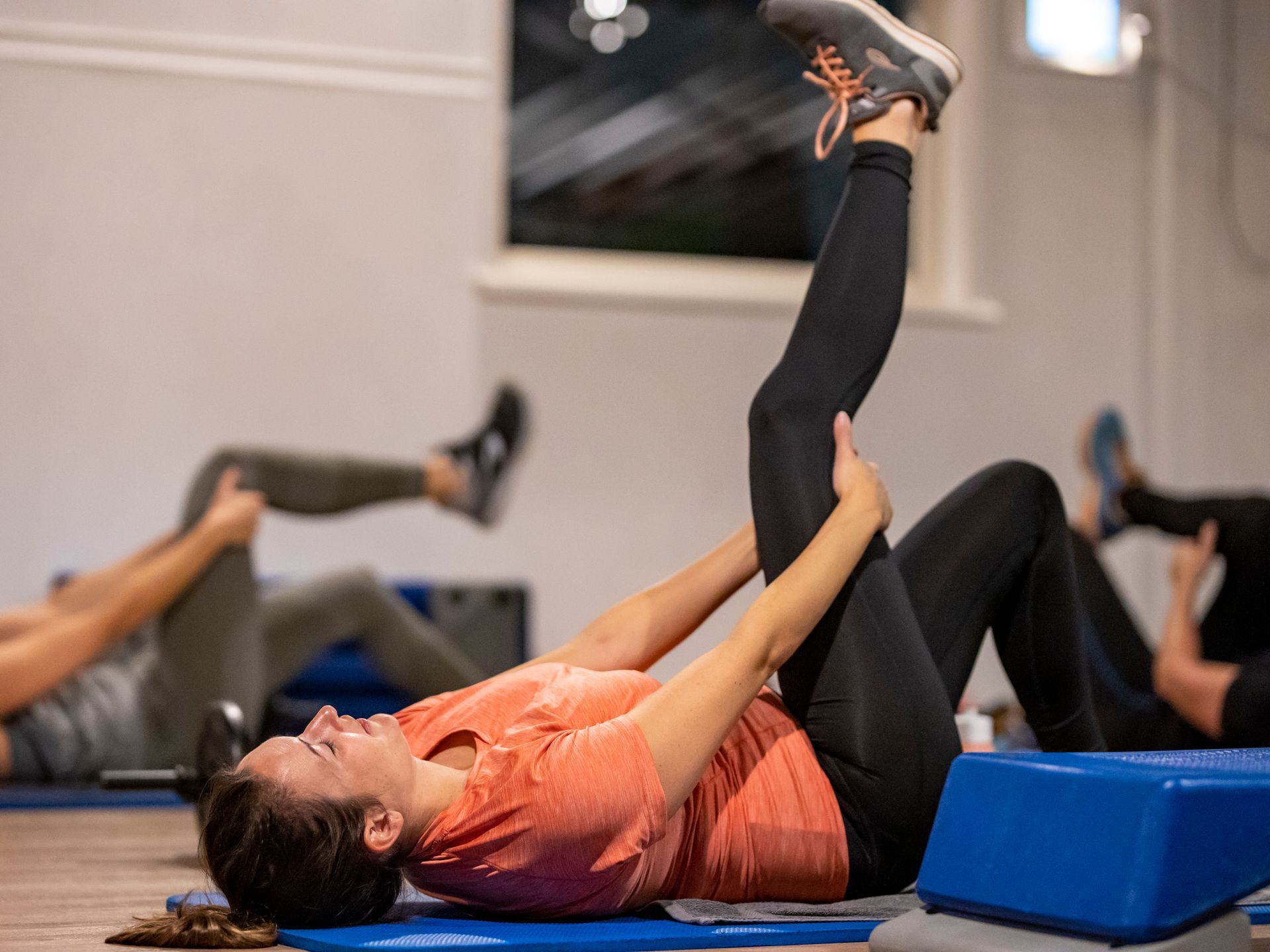 Mensen doen beenstrekoefeningen op matten in een sportschool. Een vrouw in een oranje shirt en zwarte legging strekt haar been.