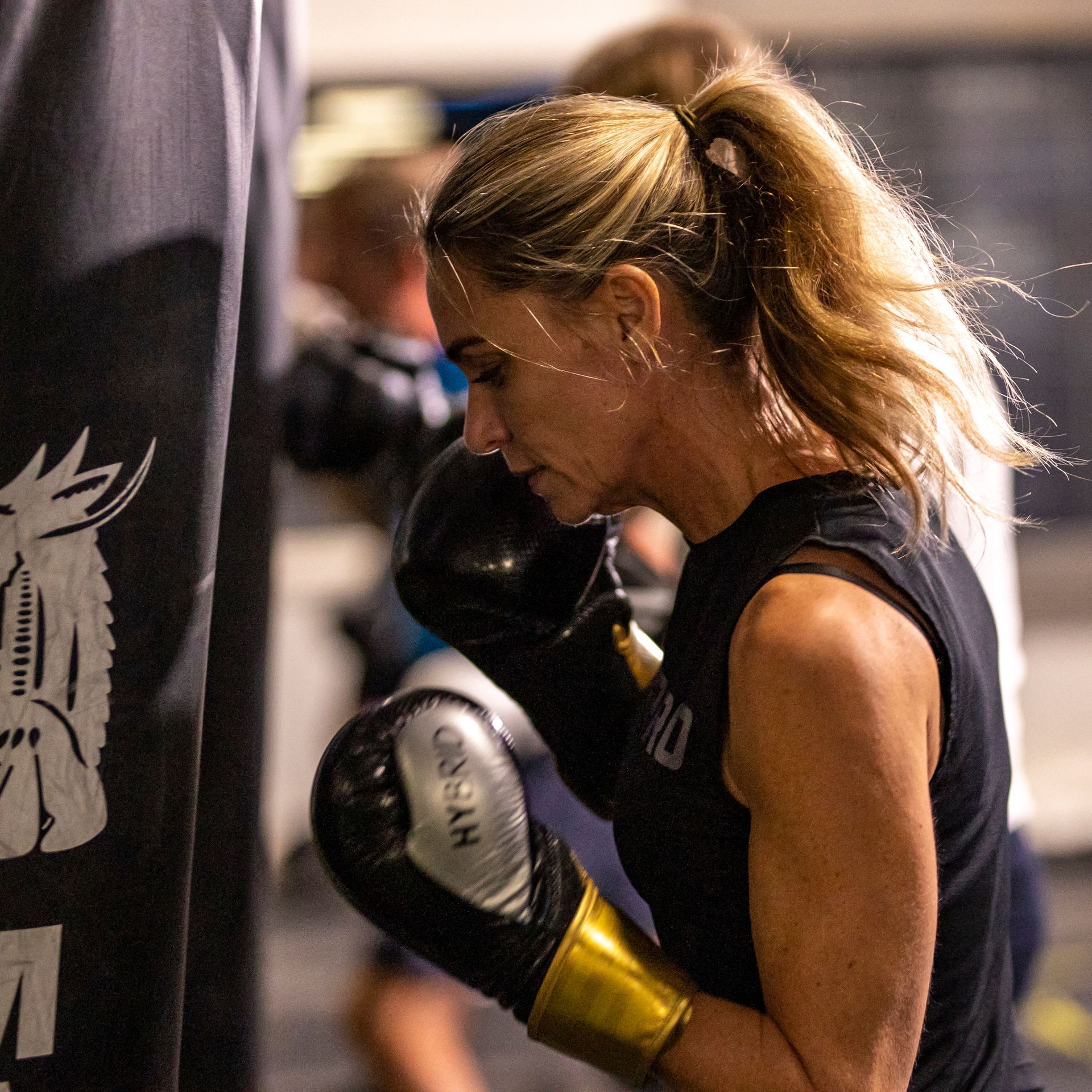 Een vrouw met bokshandschoenen slaat tegen een boksbal in een sportschool. Ze heeft blond haar in een paardenstaart en is geconcentreerd.