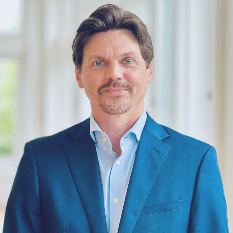 Man in blue suit jacket and white shirt, standing indoors against a bright blurred background.