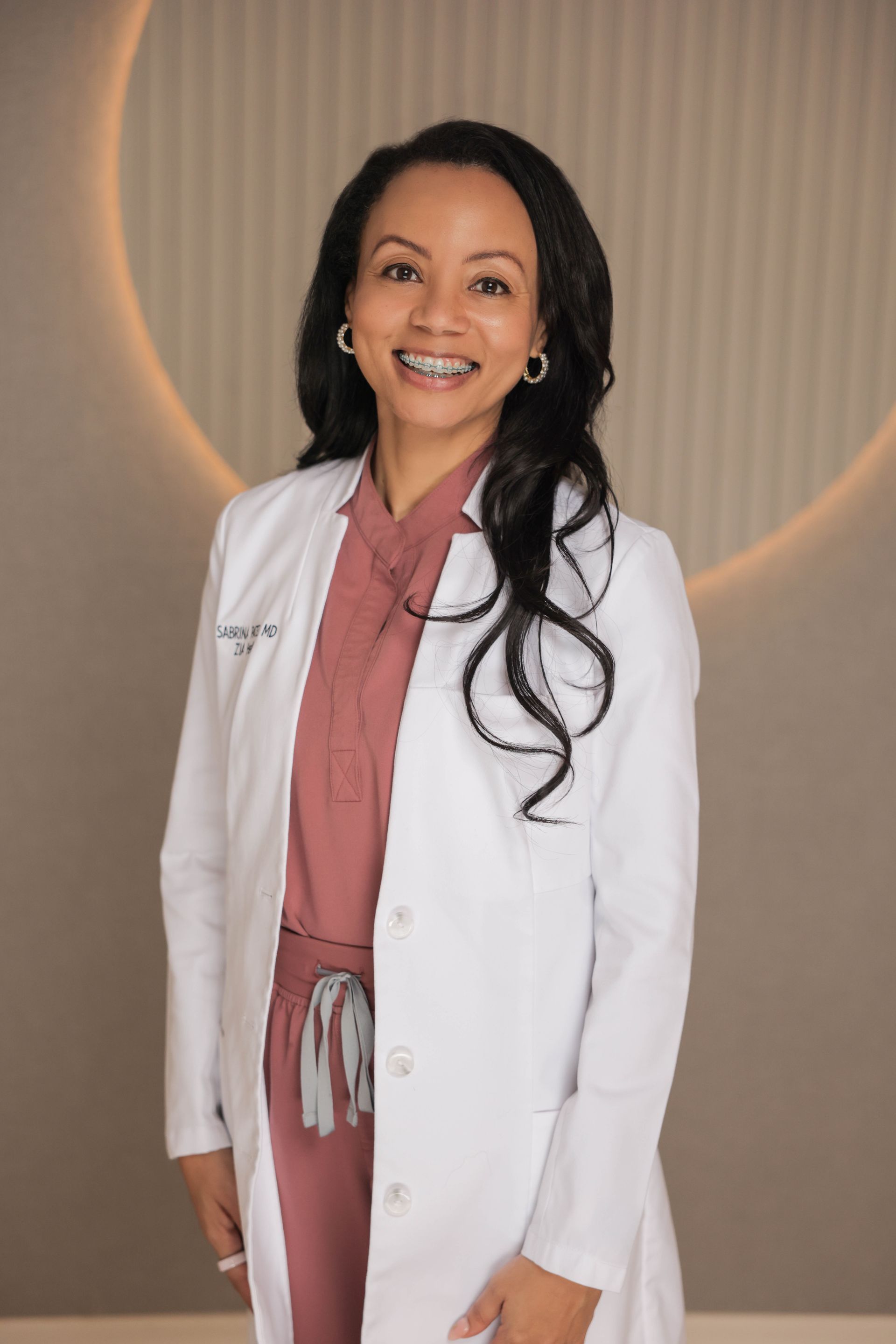 Smiling doctor in white coat and pink scrubs standing against a neutral backdrop