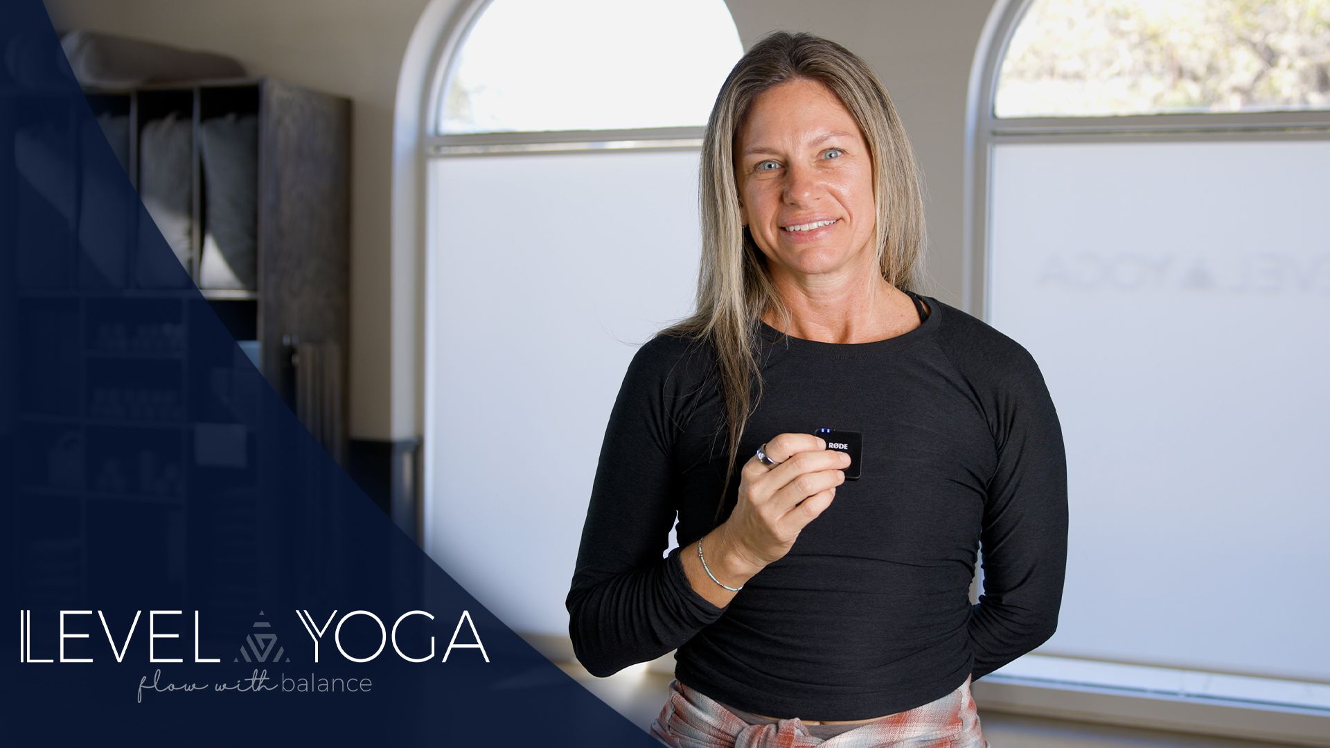 Woman in a yoga studio holds up a finger. Dark shirt, light pants, window background, and 