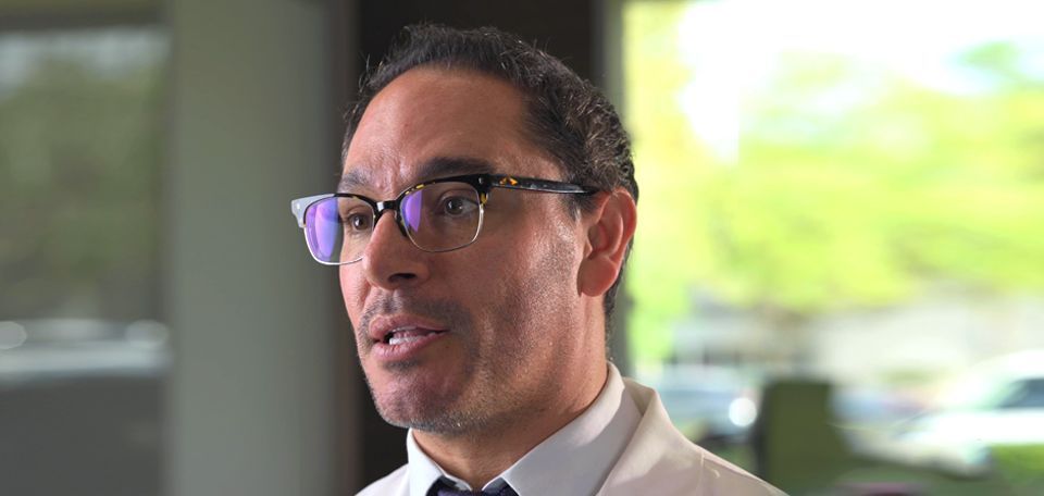 A man wearing glasses and a white coat is talking to someone.