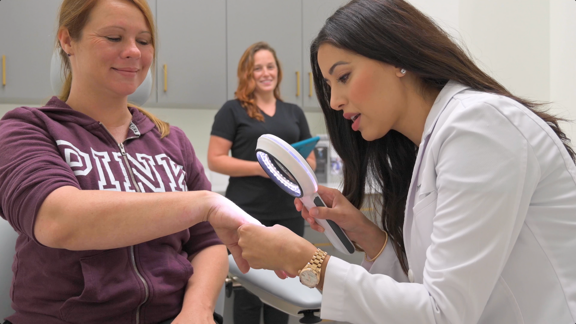 Woman examines another woman's arm with a magnifying glass in a clinical setting; one smiles in the background.