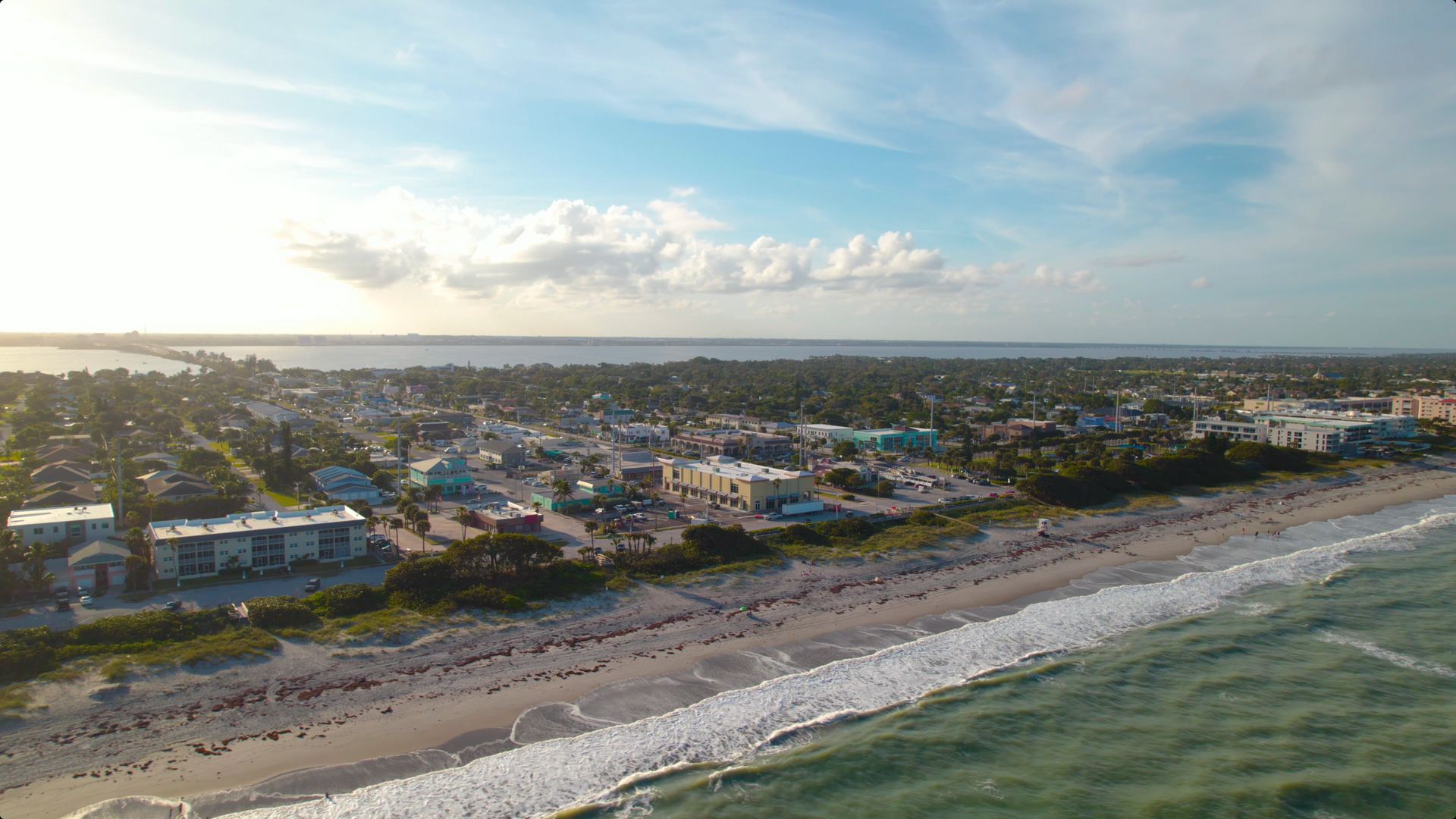 Aerial view of beach, town, and waterway under partly cloudy sky.