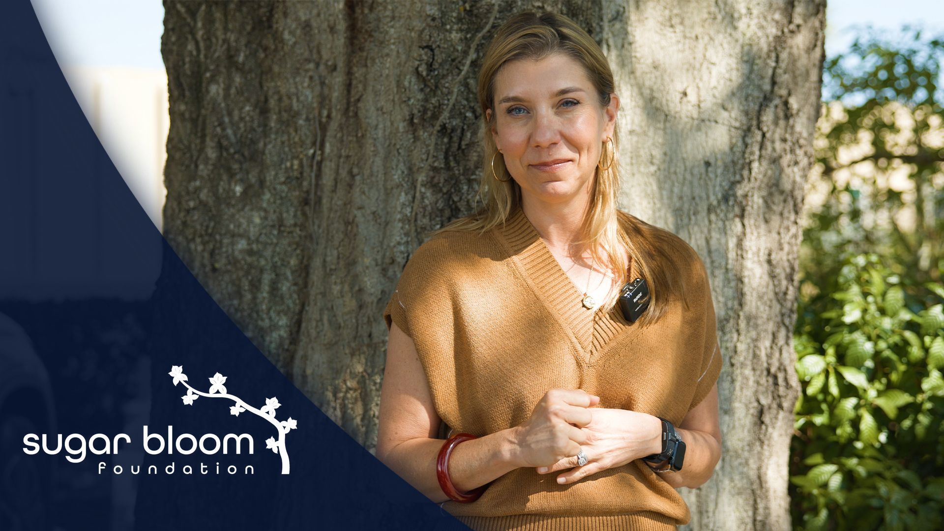Woman in brown top stands by a tree; Sugar Bloom Foundation logo in corner.