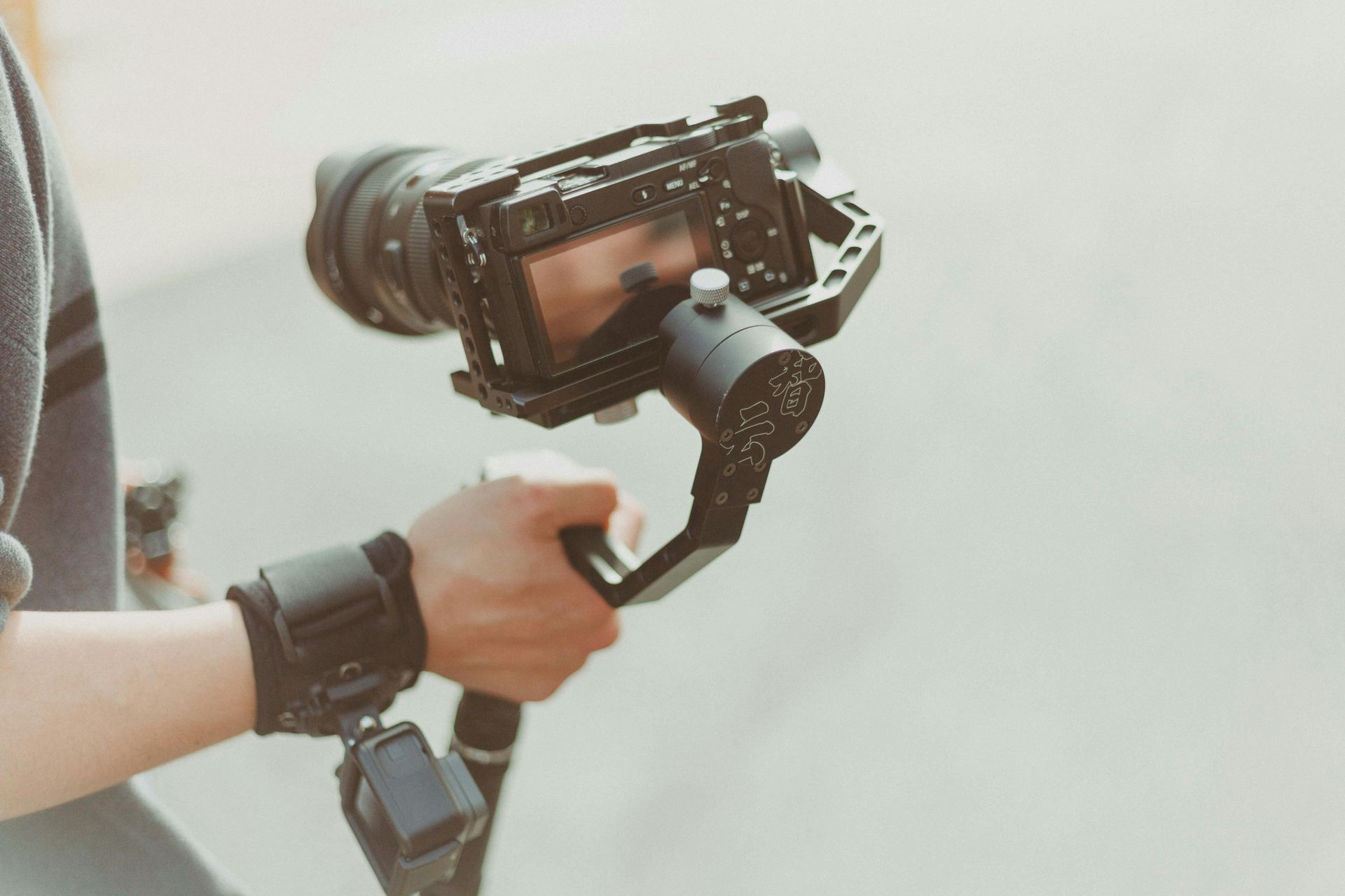 Person holding a camera mounted on a gimbal. The camera is black, with a large lens.  Gray background.
