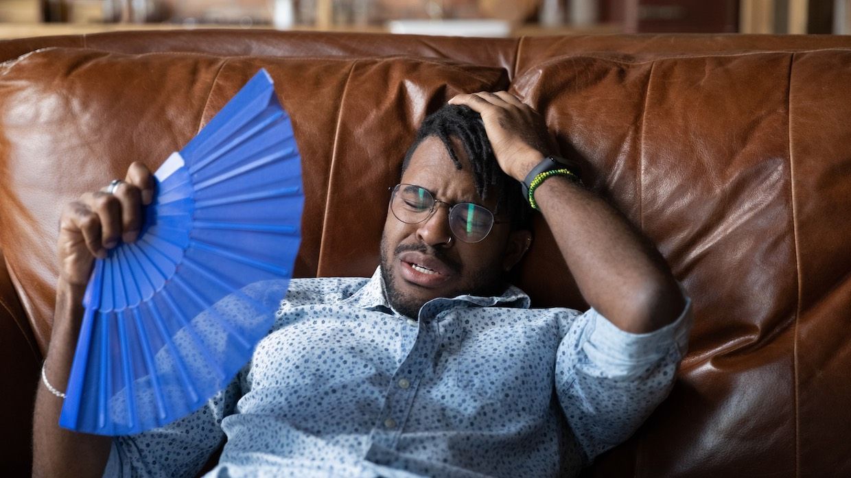 Man on brown couch, using a blue fan, looking distressed.