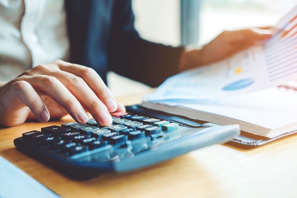 Person's hands using a calculator, with paperwork and charts in the background on a desk.