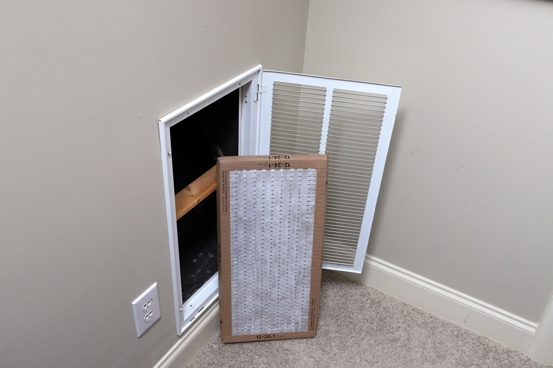 A dusty air filter, open access panel, and a metal grate in a corner of a room, next to a power outlet.