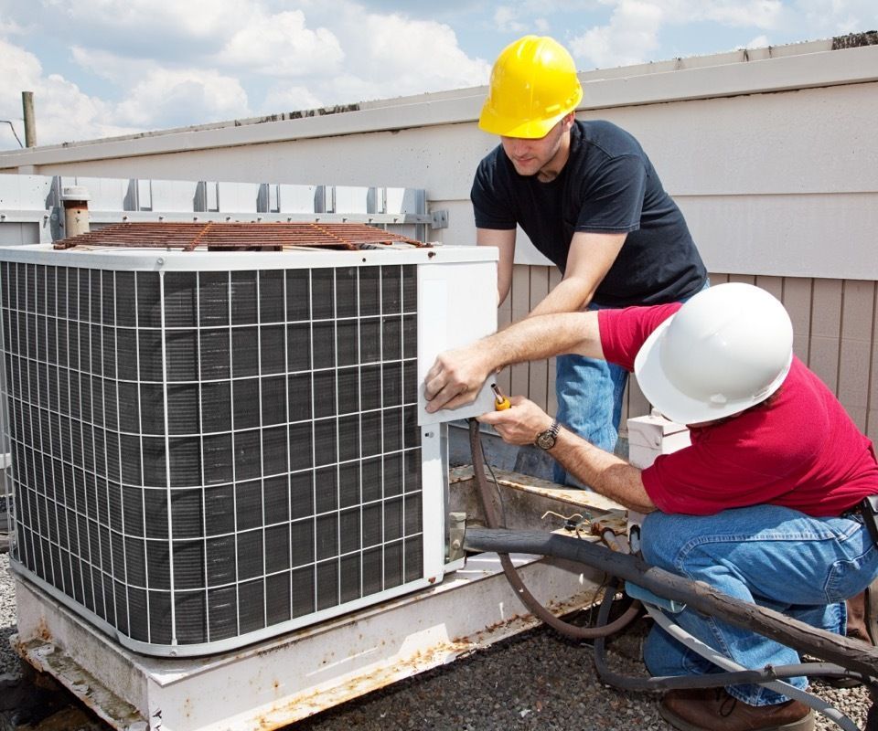 Two HVAC technicians in hard hats work on an air conditioning unit on a rooftop.