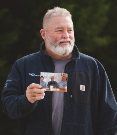 Man holding a photo of three men; he wears a blue fleece with a gray shirt. Outdoors, smiling.