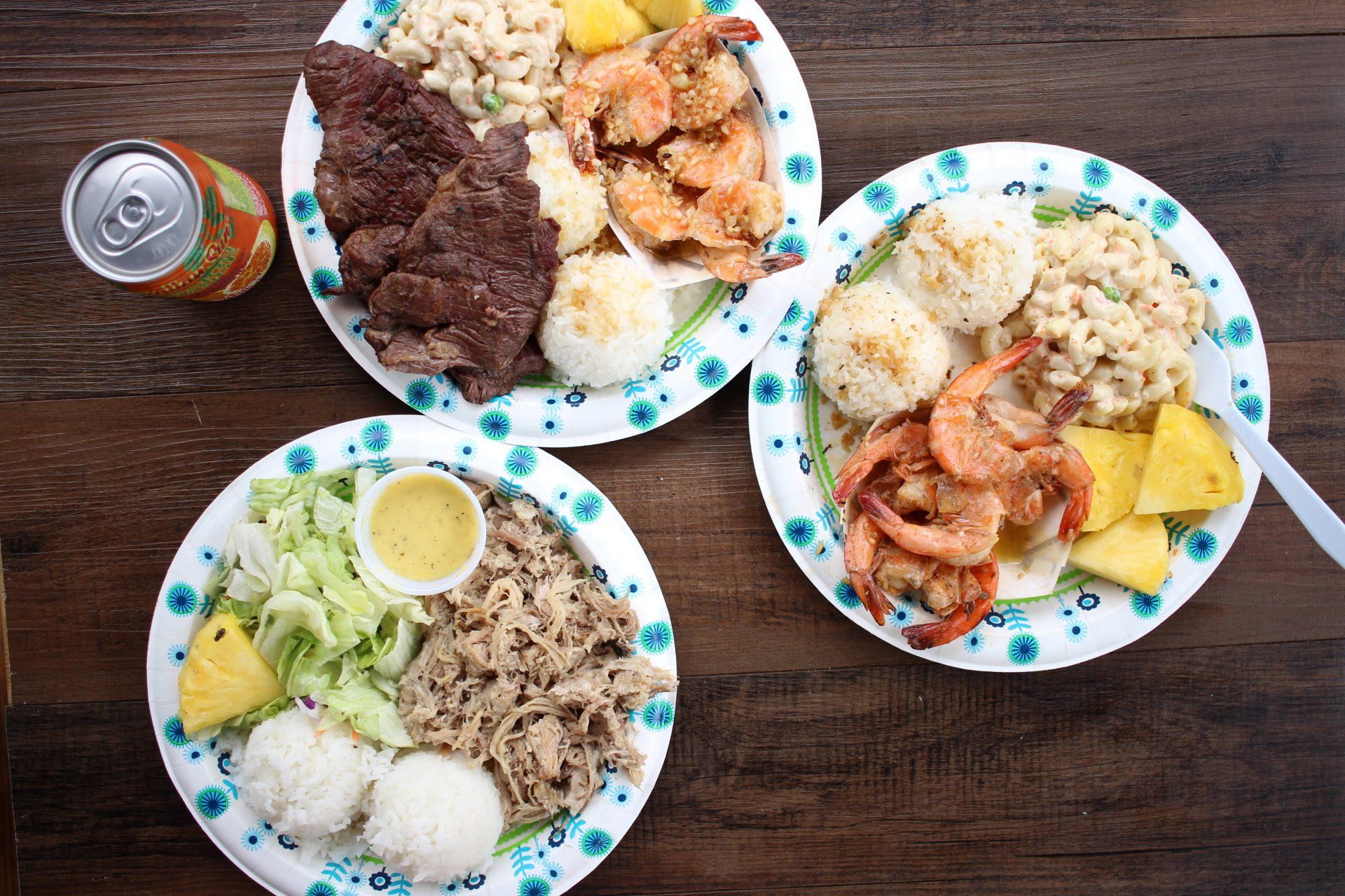 Three plates of Hawaiian plate lunches with various meats, rice, macaroni salad, and pineapple, with a canned drink.