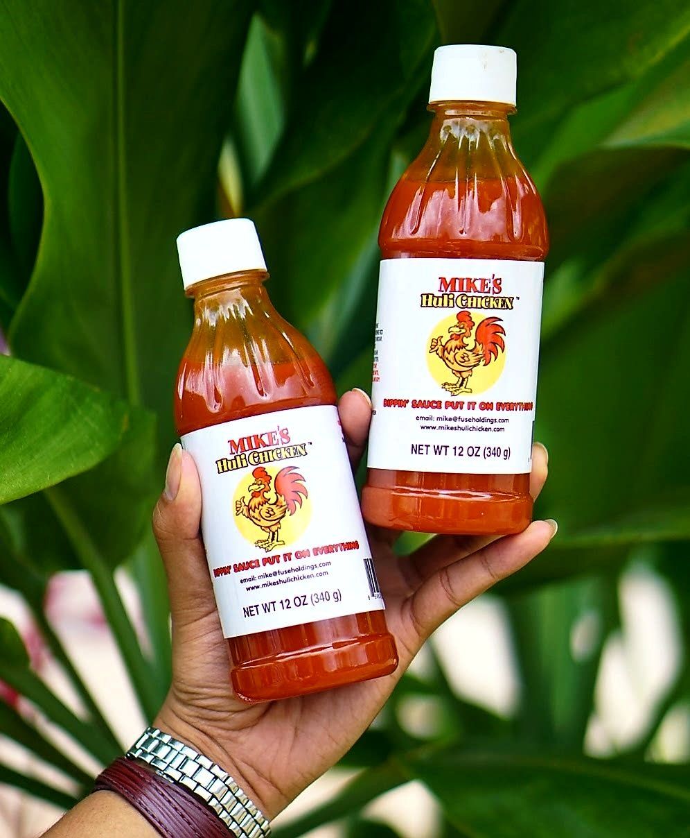 Hand holding two bottles of Mike's Hot Honey hot sauce against a background of green leaves.