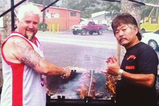 Guy Fieri grilling ribs with a man, possibly in a roadside setting; both smiling over the BBQ.