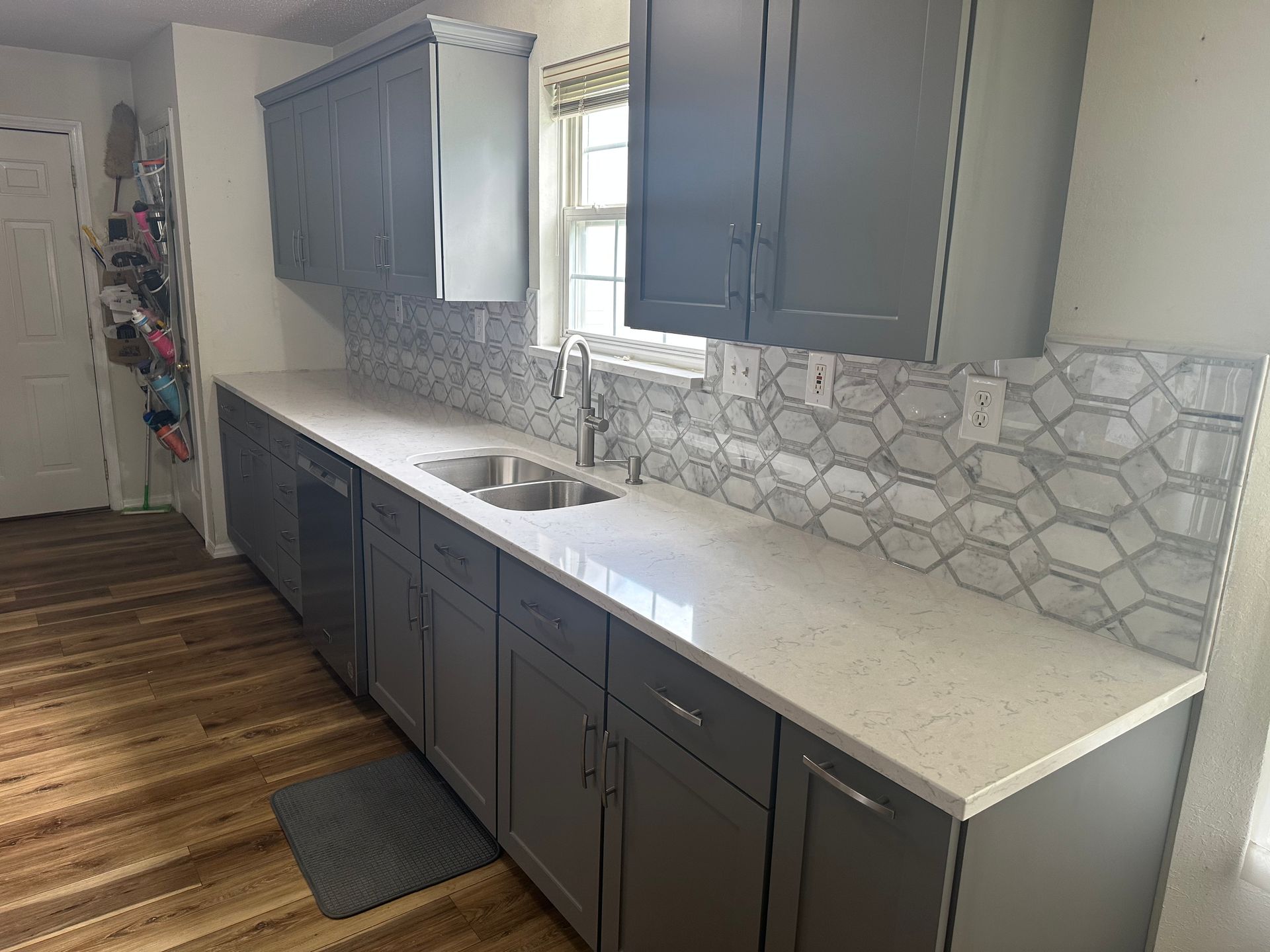 A kitchen with gray cabinets , white counter tops , a sink and a window.