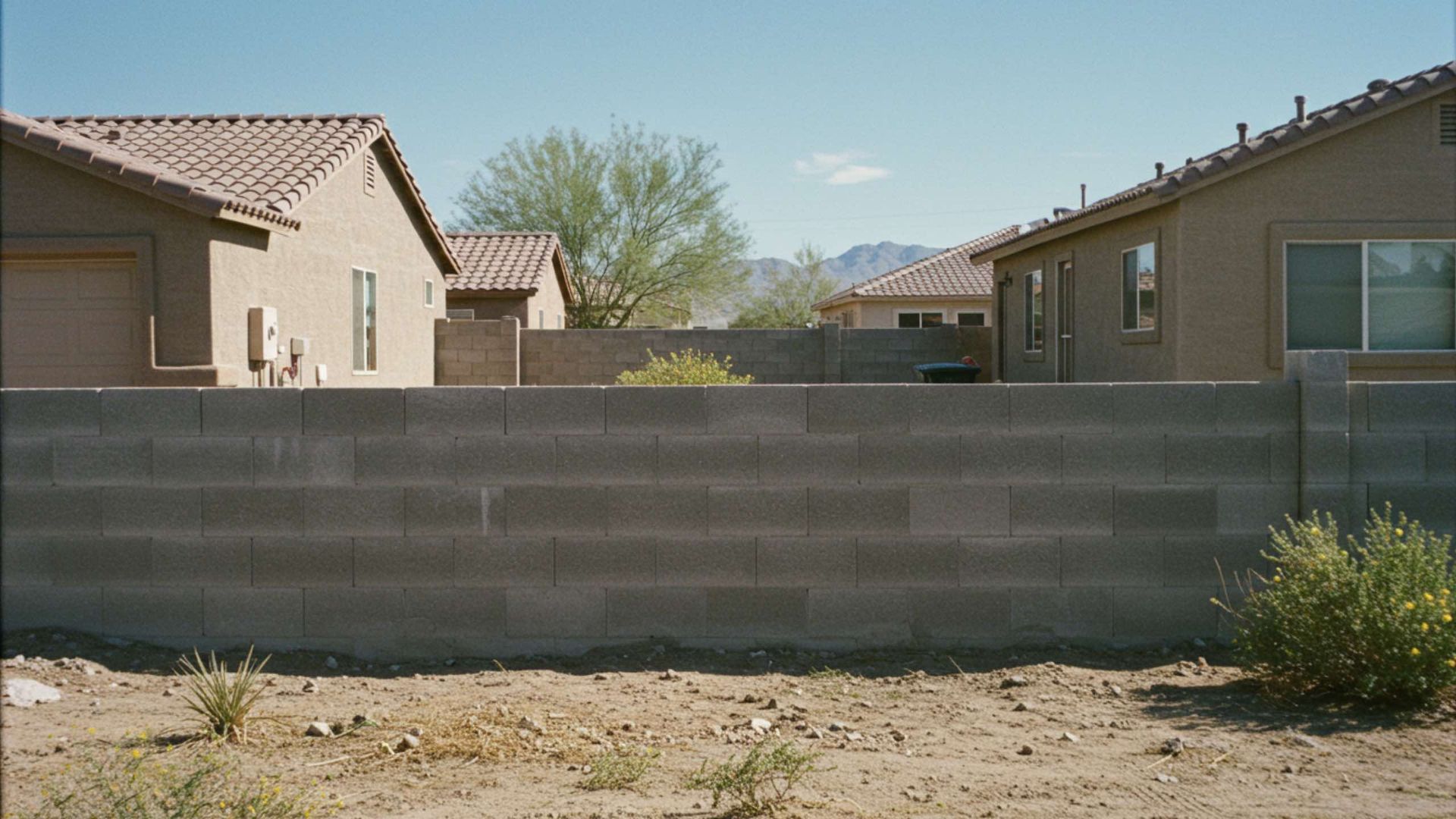 Gray cinder block wall in front of beige houses with red-tile roofs and a dry, dusty yard.