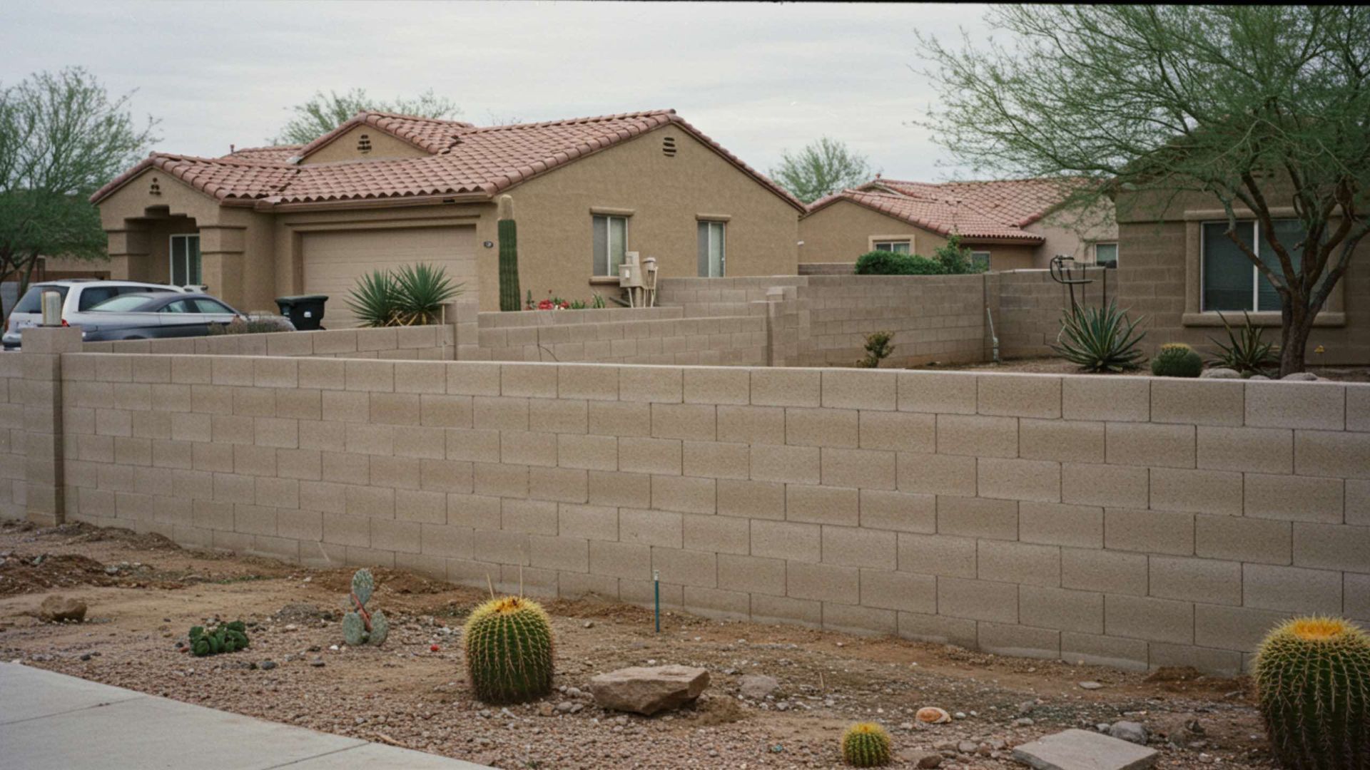 A tan block wall in front of a beige house with a red tile roof. Desert plants in front.