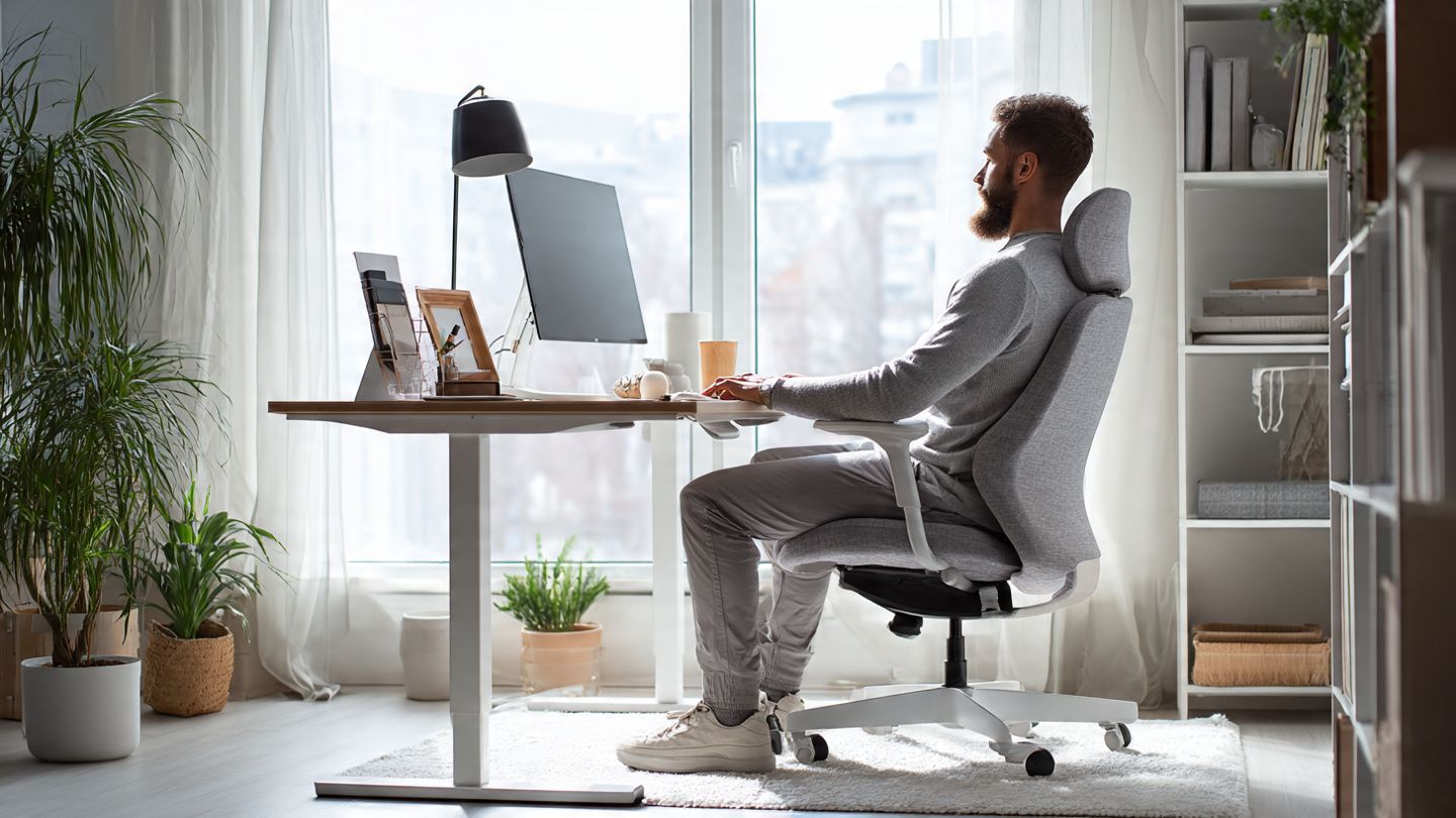 A person in a gray matching set sits at a modern desk in a sunlit home office, working on a computer.