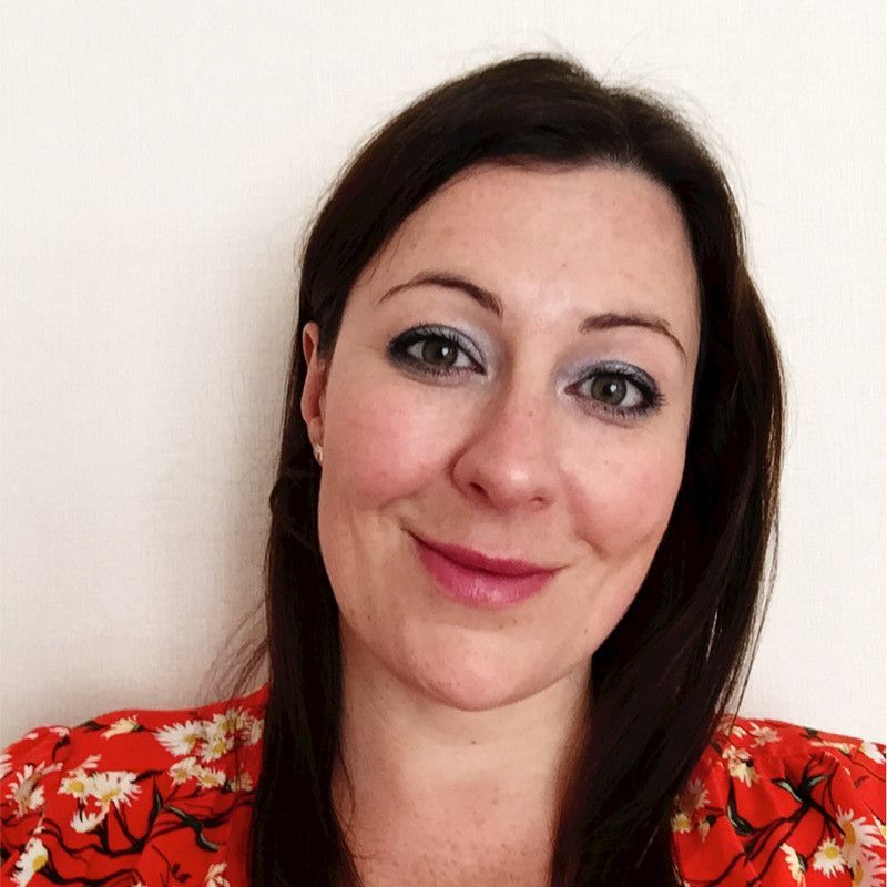 A woman with dark hair and a light smile, wearing a red floral-patterned top against a plain white background.