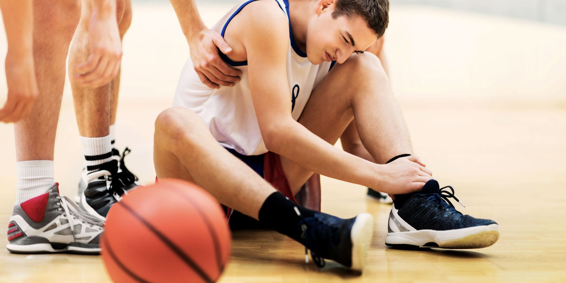 A basketball player is sitting on the floor with his ankle in pain.