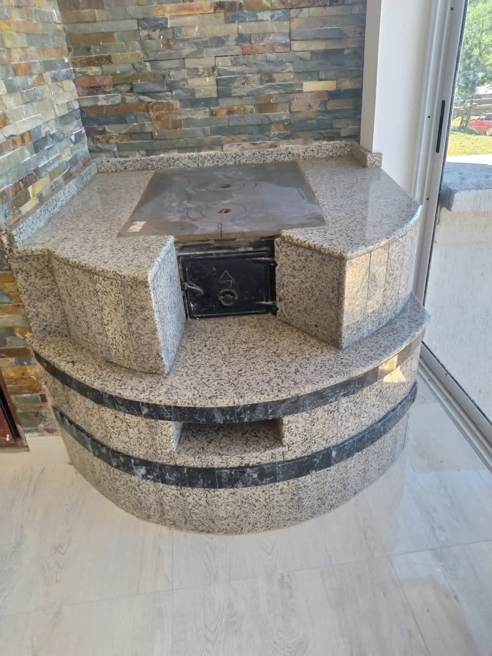 Parrilla exterior de granito con detalles negros contra una pared de piedra y puerta de vidrio.