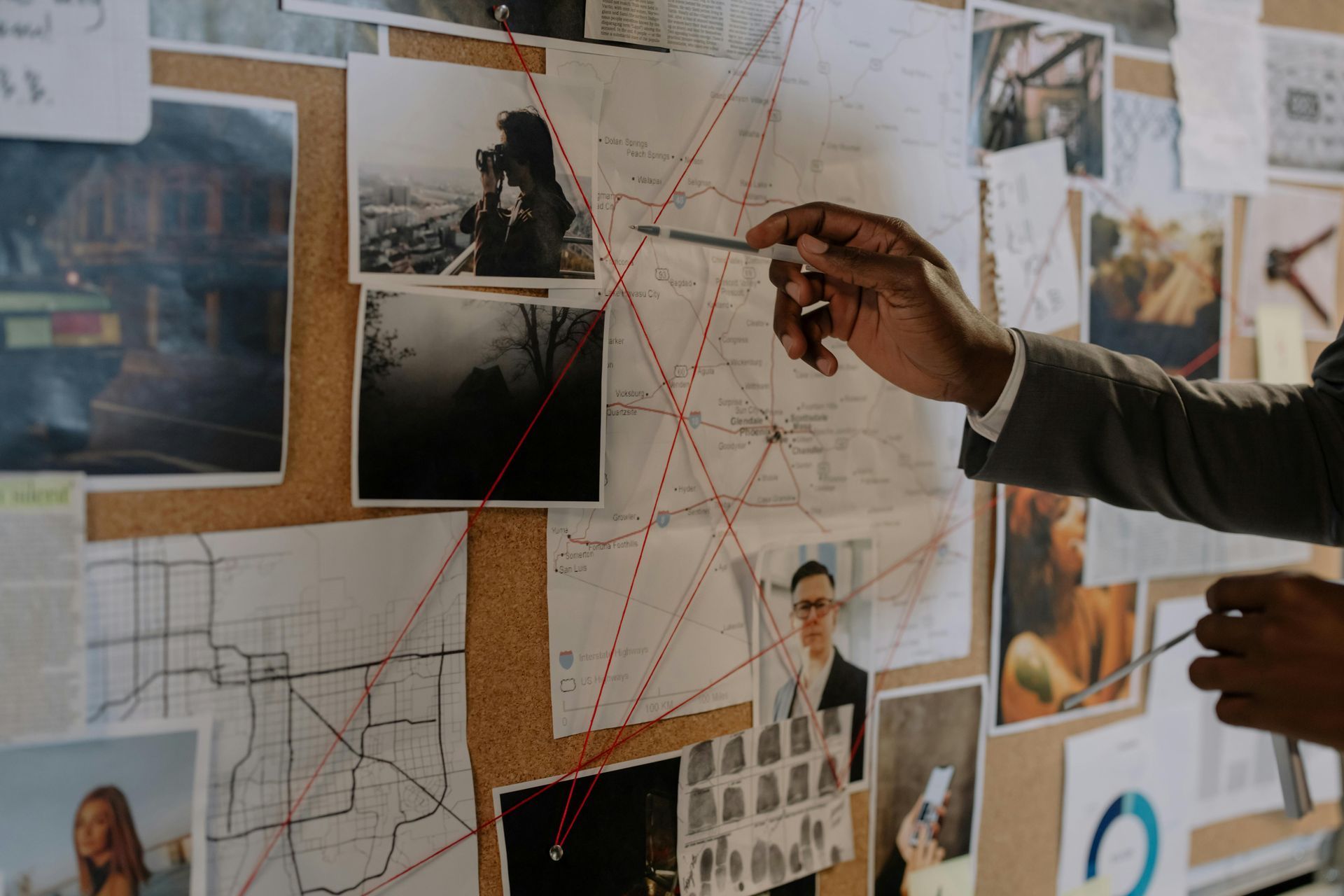 Person pointing at a corkboard covered in photos and maps, connected by red string, in an investigation setting.